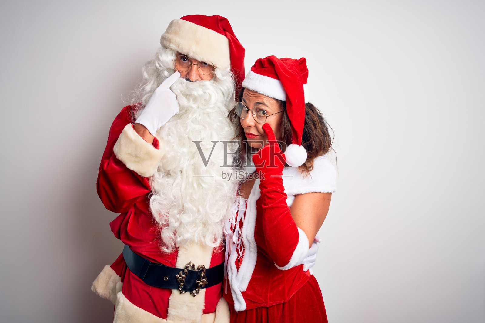 中年夫妇穿着圣诞老人服装拥抱在孤立的白色背景指向眼睛看着你的手势，可疑的表情照片摄影图片