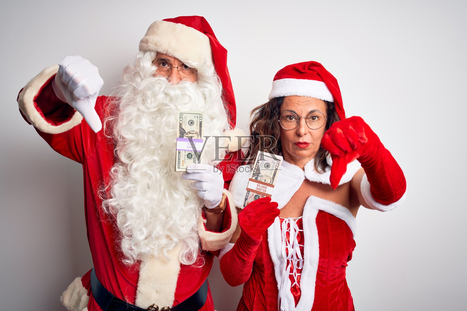 一对老年夫妇穿着圣诞老人服装，拿着美元在孤立的白色背景上，看起来不高兴和生气，用拇指向下的手势表示拒绝和否定。糟糕的表达。照片摄影图片