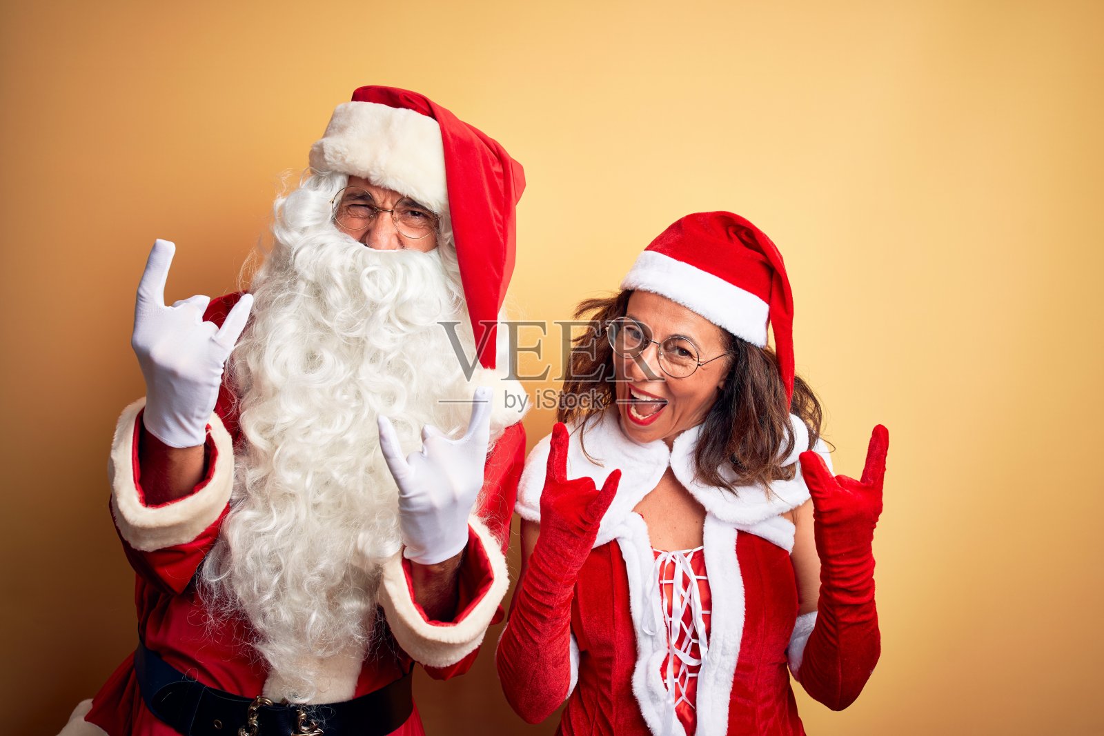 中年夫妇穿着圣诞老人服装和眼镜在孤立的黄色背景大喊与疯狂的表情做摇滚符号与手举起。音乐明星。沉重的概念。照片摄影图片