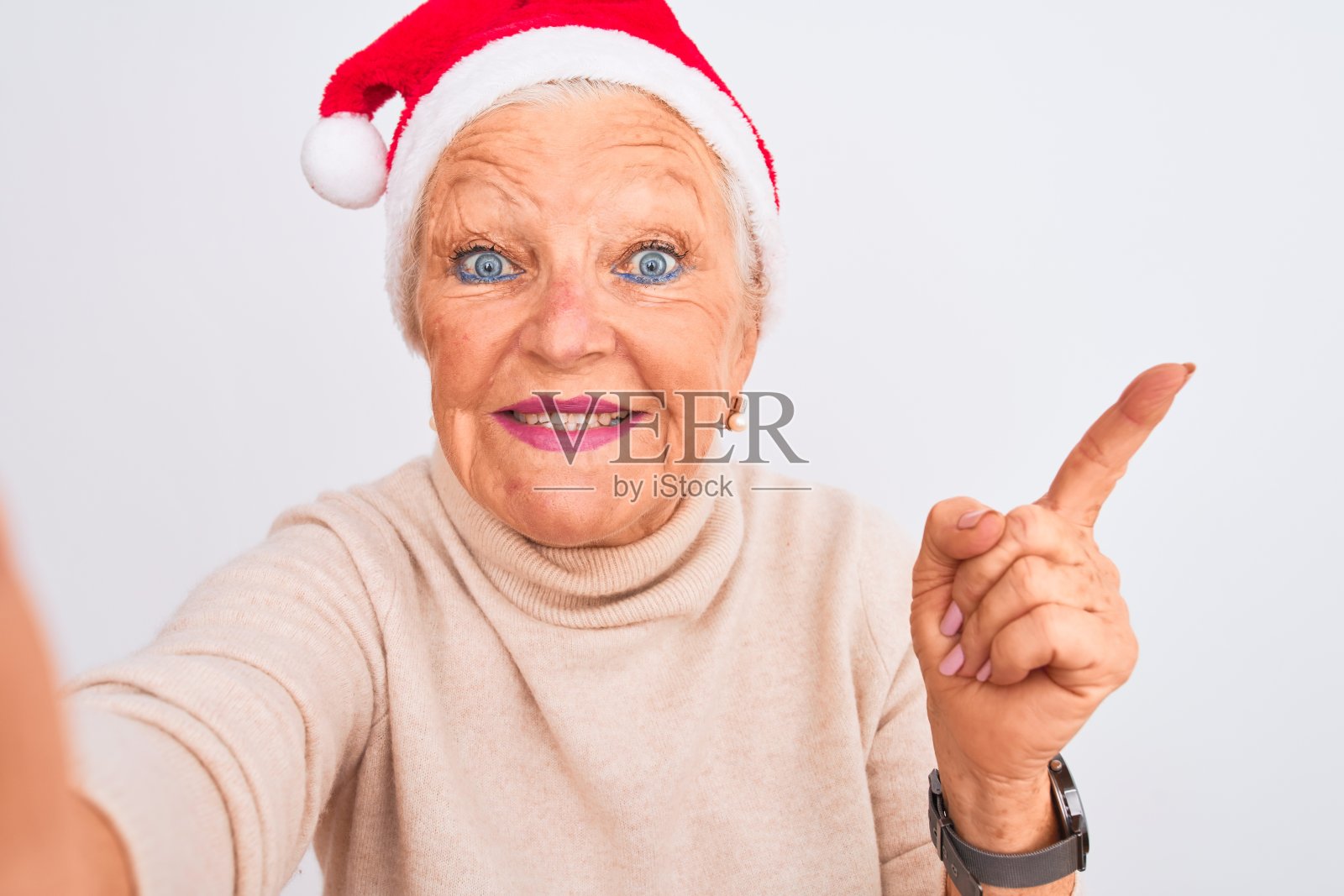 灰白头发的女人戴着圣诞老人的帽子在孤立的白色背景上自拍，非常开心地用手和手指指向一边照片摄影图片