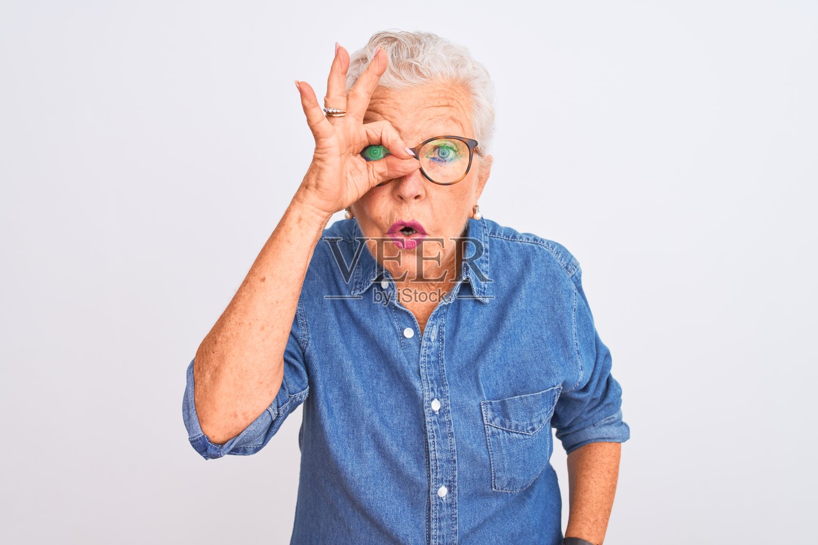 资深灰头发妇女穿着牛仔衬衫和眼镜在孤立的白色背景做ok手势震惊与惊讶的脸，眼睛透过手指。不相信的表情。照片摄影图片
