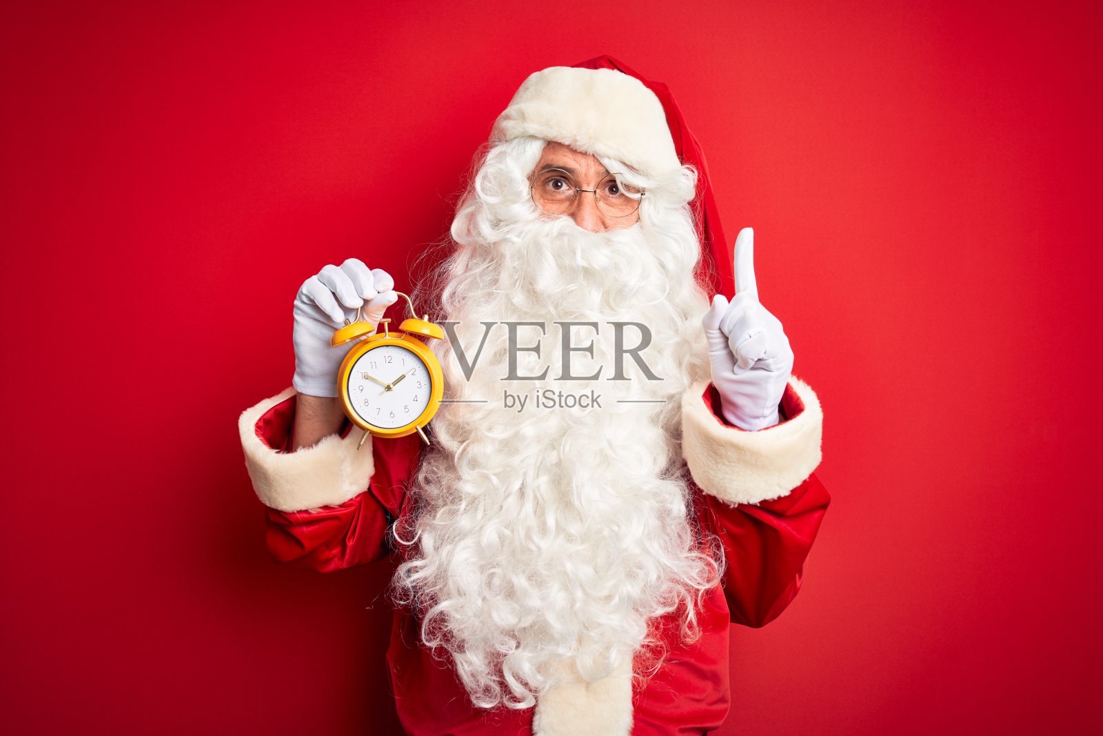 一名身穿圣诞老人服装的老人手持闹钟在孤立的红色背景下惊讶地提出一个想法或问题，用幸福的脸指着手指，第一个照片摄影图片