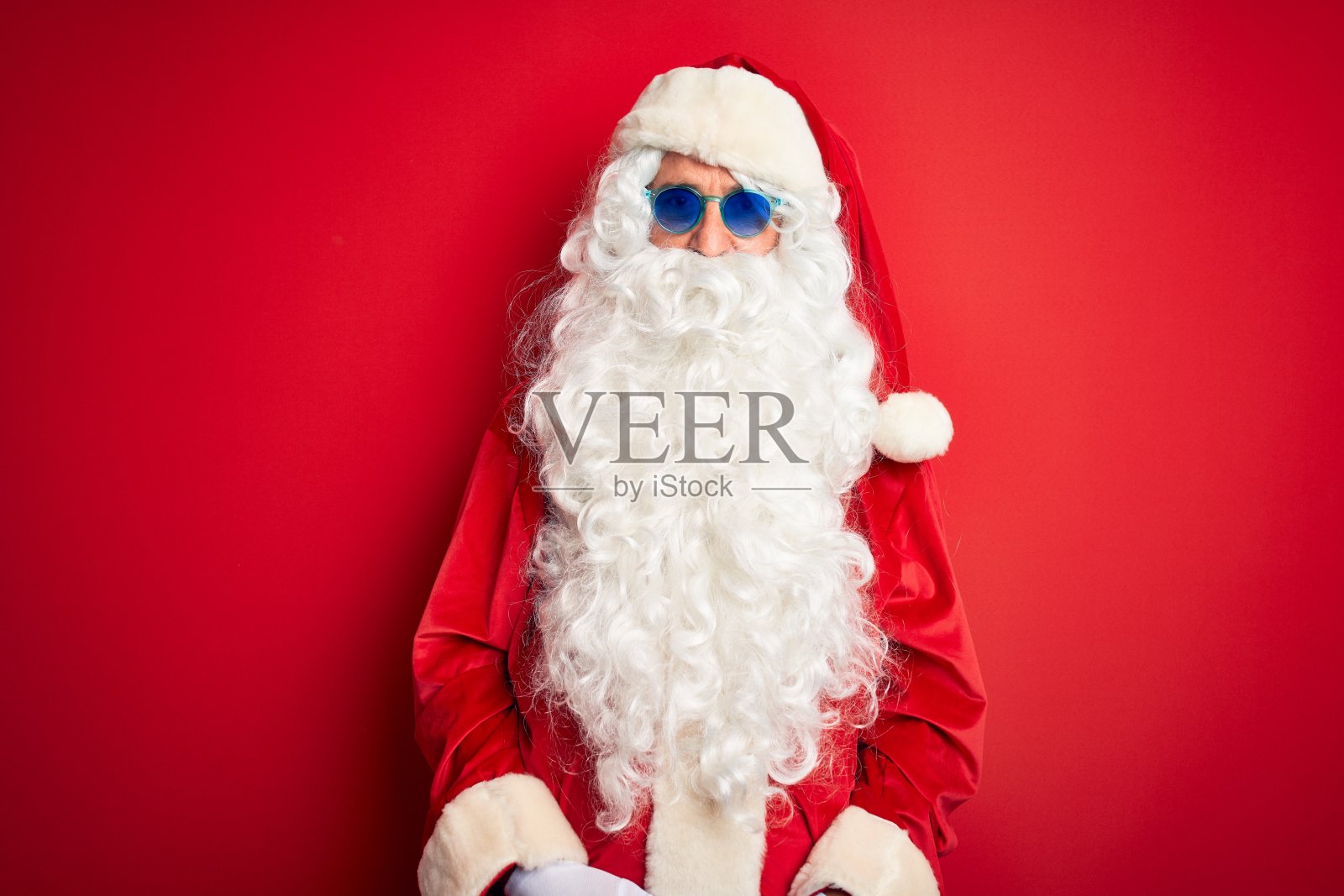 中年男子穿着圣诞老人服装和太阳眼镜在孤立的红色背景与严肃的表情在脸上。简单自然的看相机。照片摄影图片