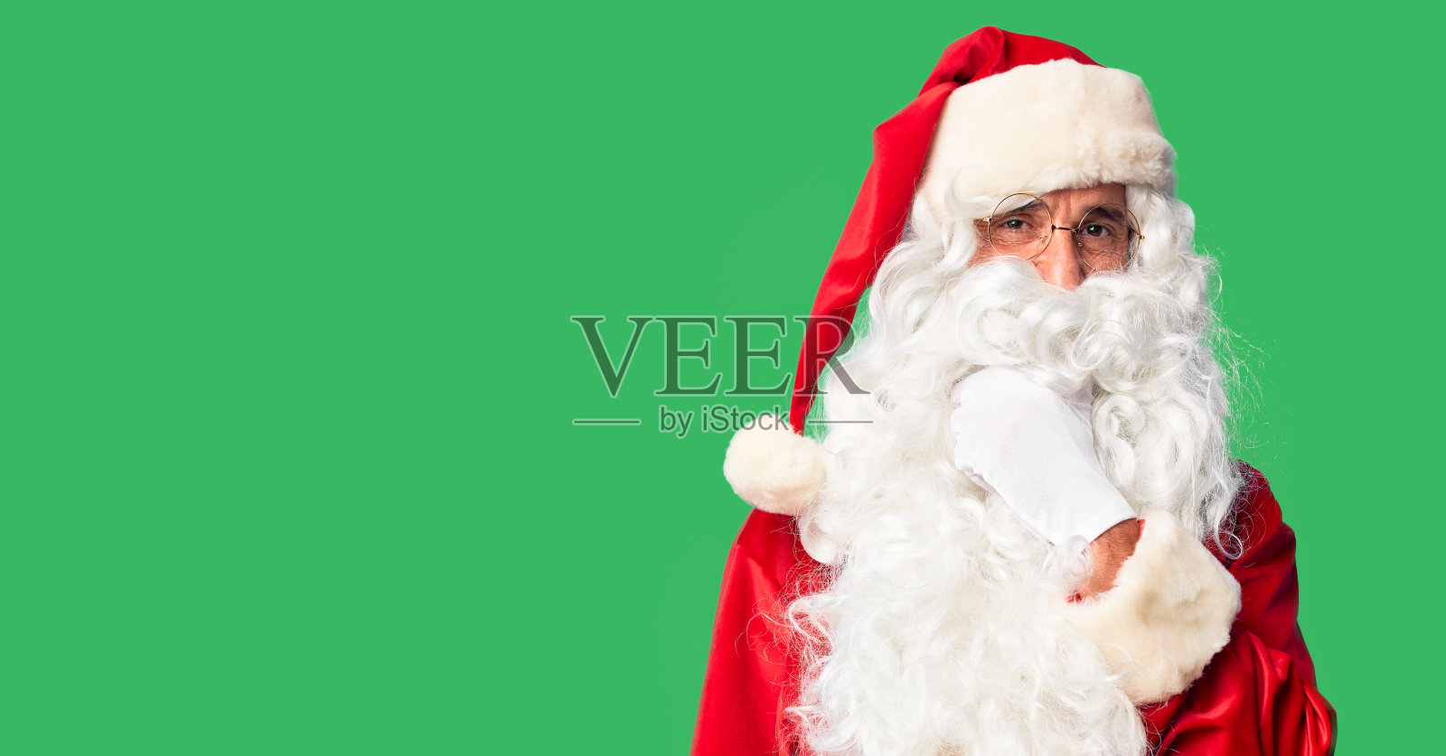 英俊的中年男子穿着圣诞老人的服装和胡子站在镜头前看起来很自信，双臂交叉，手举在下巴上微笑。积极的思考。照片摄影图片