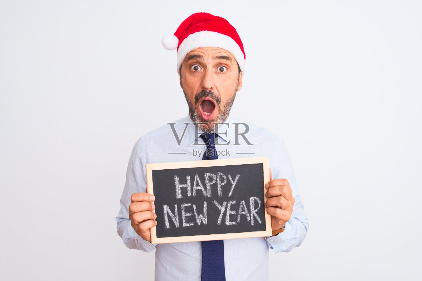 高级商人戴着圣诞帽，举着黑板越过孤立的白色背景吓得目瞪口呆，脸上带着惊恐和兴奋的恐惧表情照片摄影图片