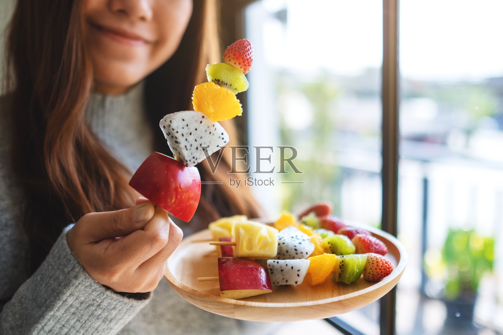 一个亚洲妇女拿着并吃着一个新鲜的混合水果串照片摄影图片