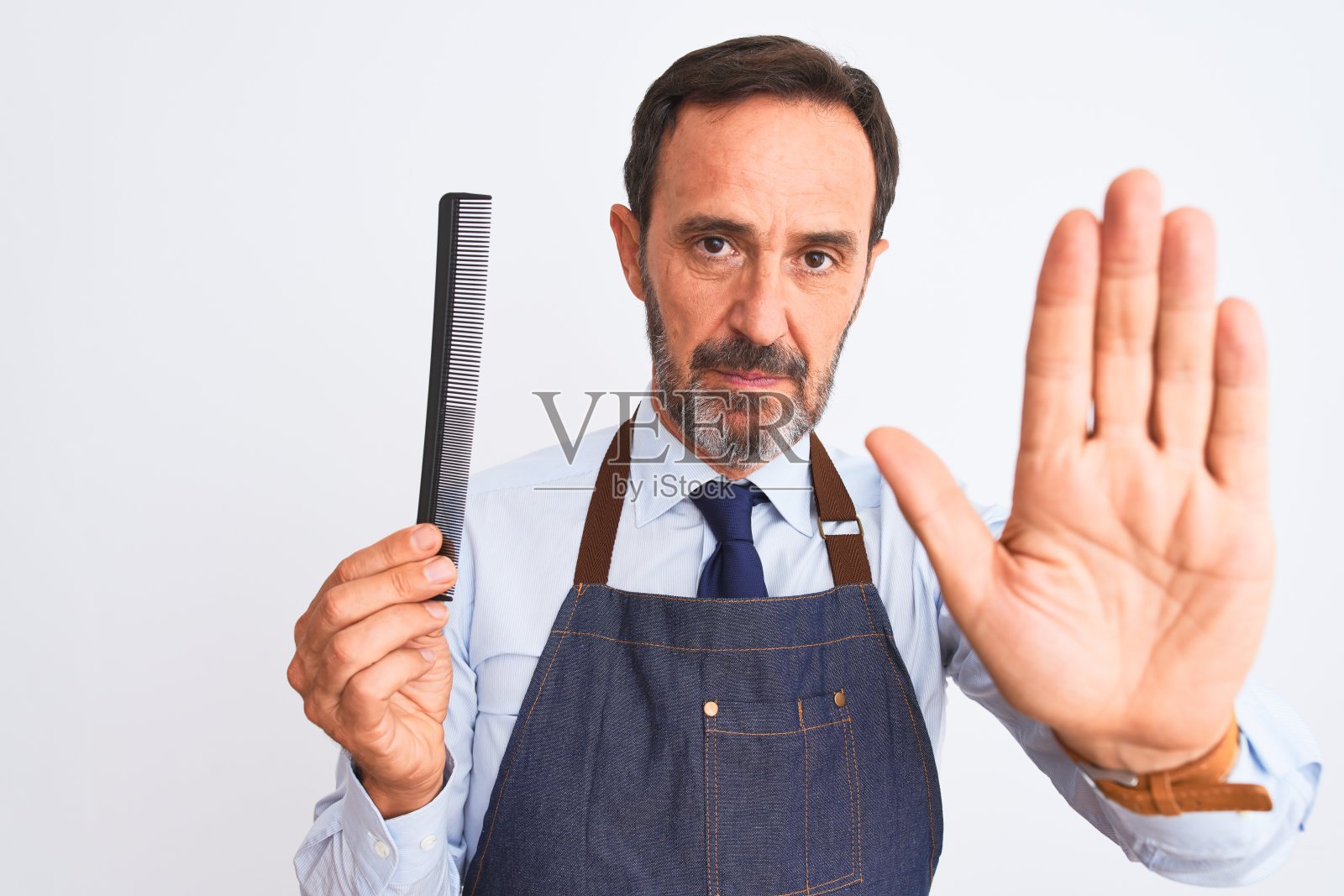 中年理发师穿着围裙拿着头发梳在孤立的白色背景与开放的手做停止标志与严肃和自信的表情，防御手势照片摄影图片