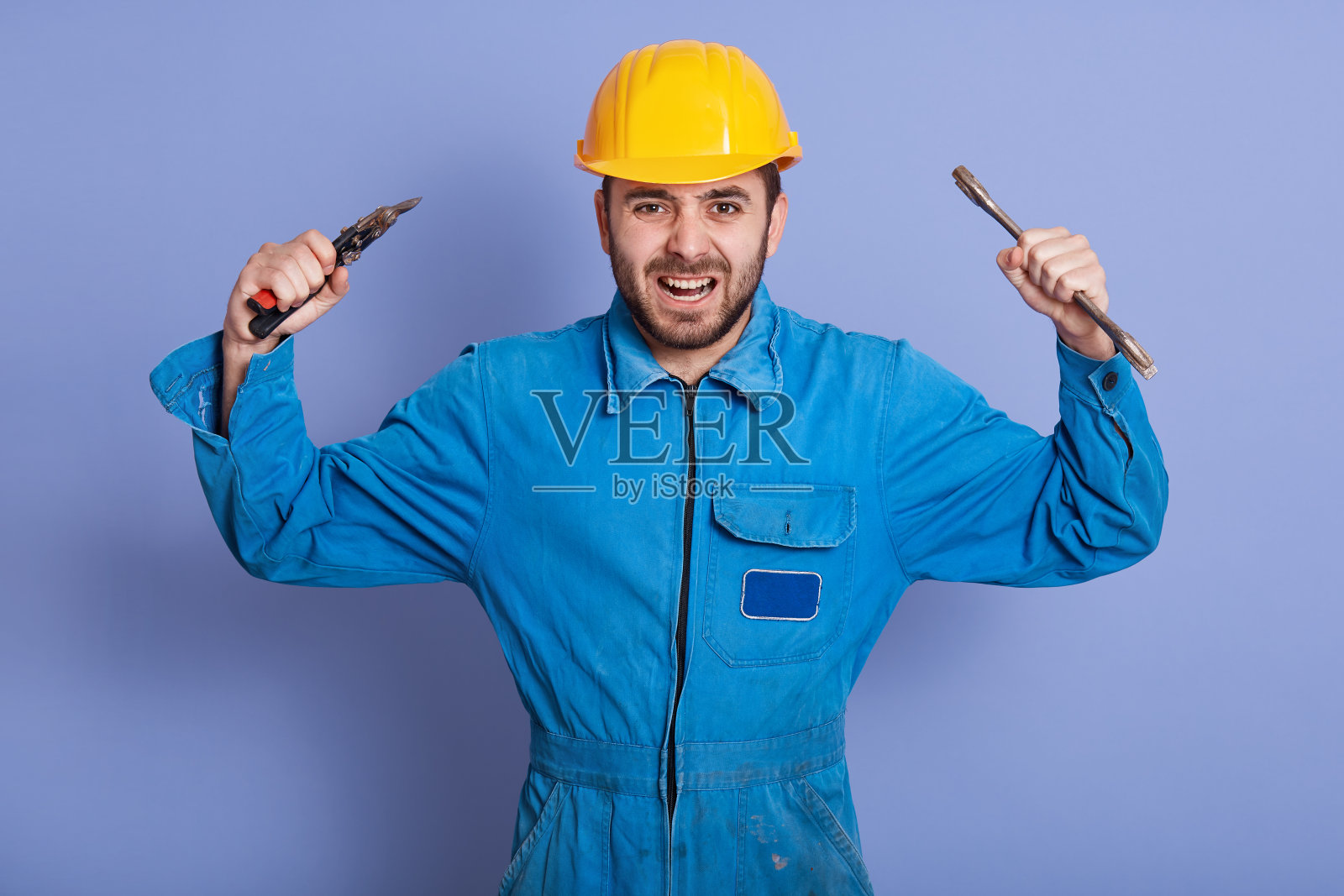 近距离的肖像，愤怒的建筑工人工程师穿着黄色头盔和蓝色制服，保持双手向上，大喊什么与愤怒，胡子的家伙拿扳手工具与双手。照片摄影图片