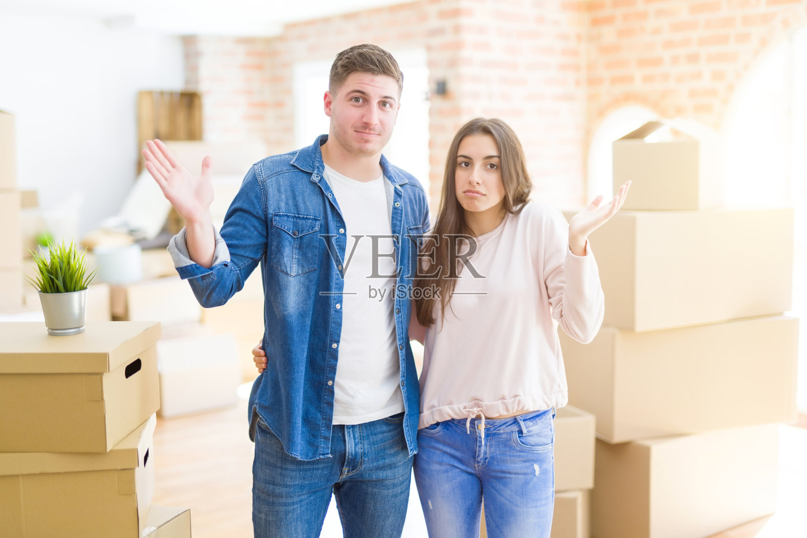 美丽的年轻夫妇搬到一个新房子，毫无头绪和困惑的表情与胳膊和手举起。怀疑的概念。照片摄影图片