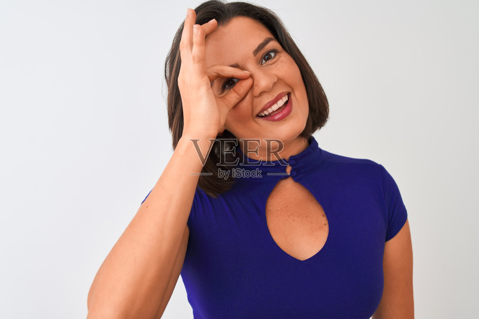 年轻美丽的女人穿着蓝色优雅的t恤站在孤立的白色背景与快乐的脸微笑做ok手势与手在眼睛看通过手指照片摄影图片