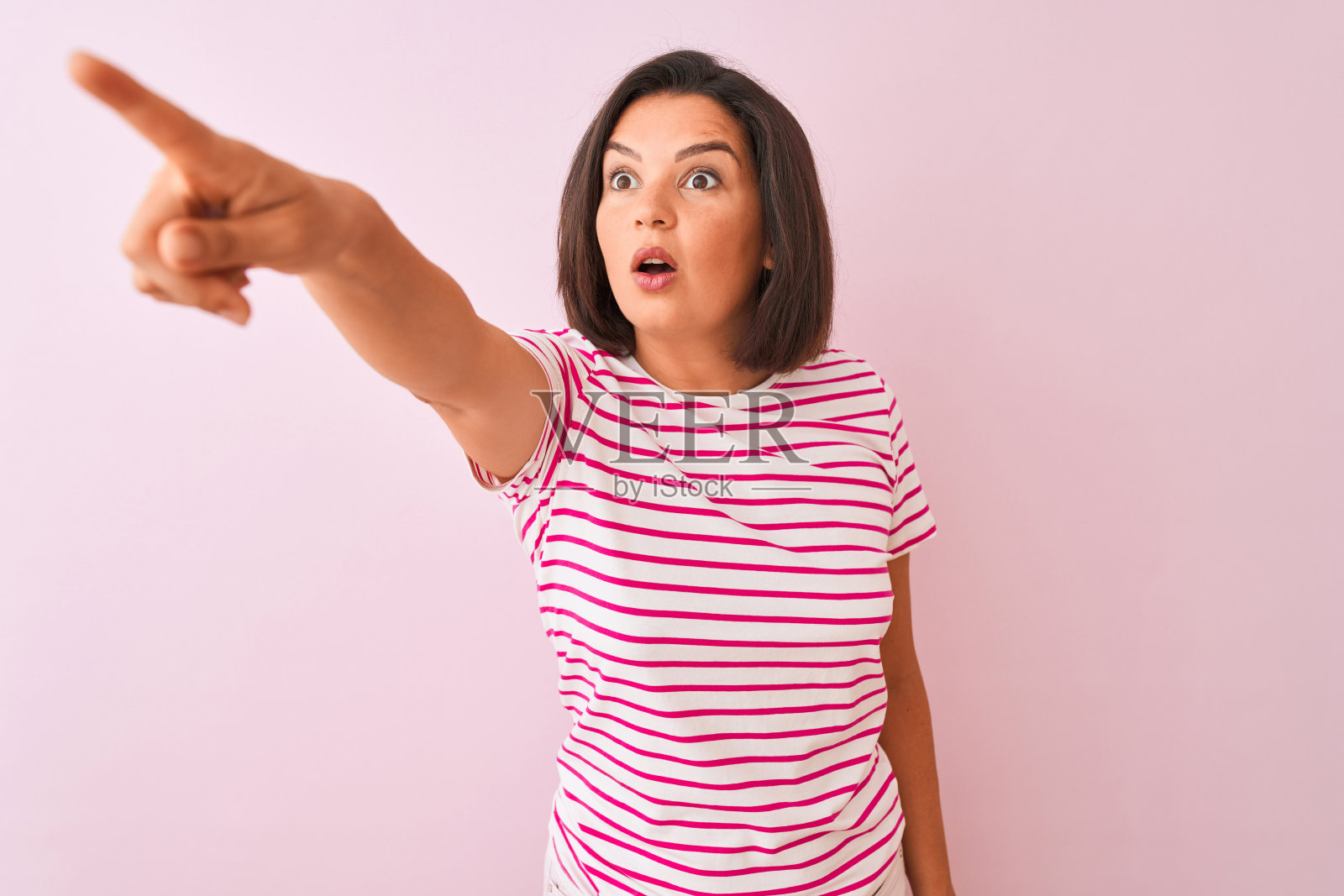 年轻漂亮的女子穿着条纹t恤站在孤立的粉红色背景上，用手指惊讶地指着前方，张着嘴惊讶地表情，前方有东西照片摄影图片