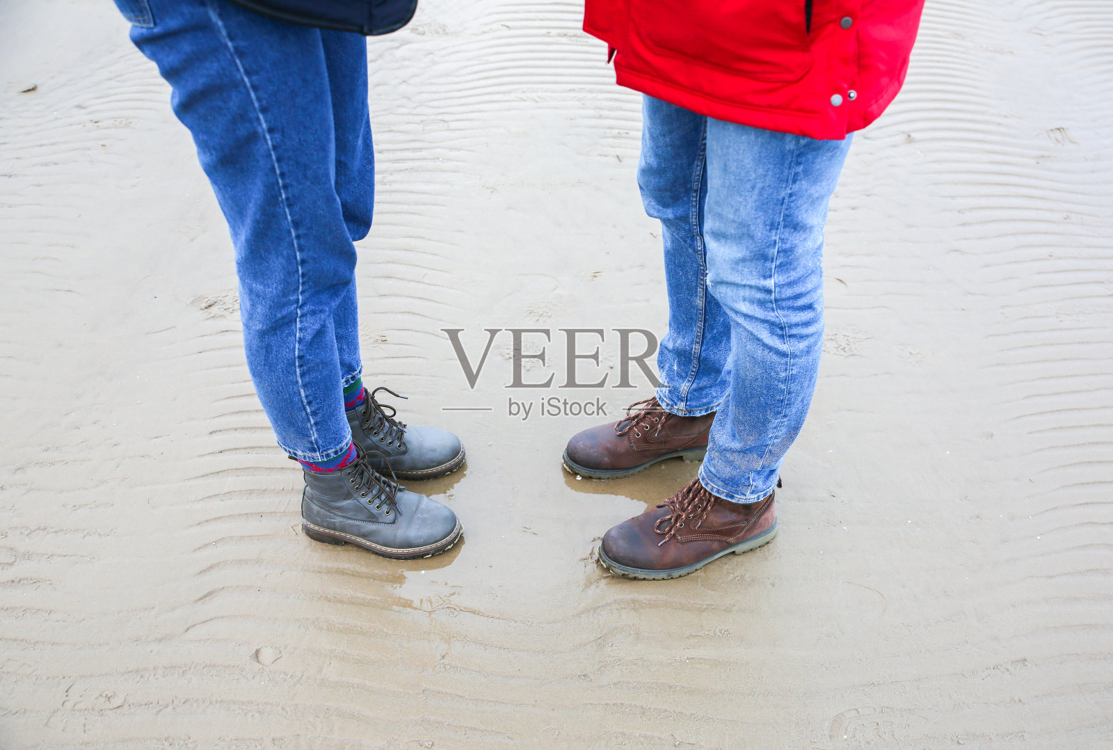 两个潮人站在冰冷的海滩上。拥抱、牵手的情侣。海边的爱情故事。冬天在海上。时髦的靴子在沙滩上。穿红夹克的男人。照片摄影图片