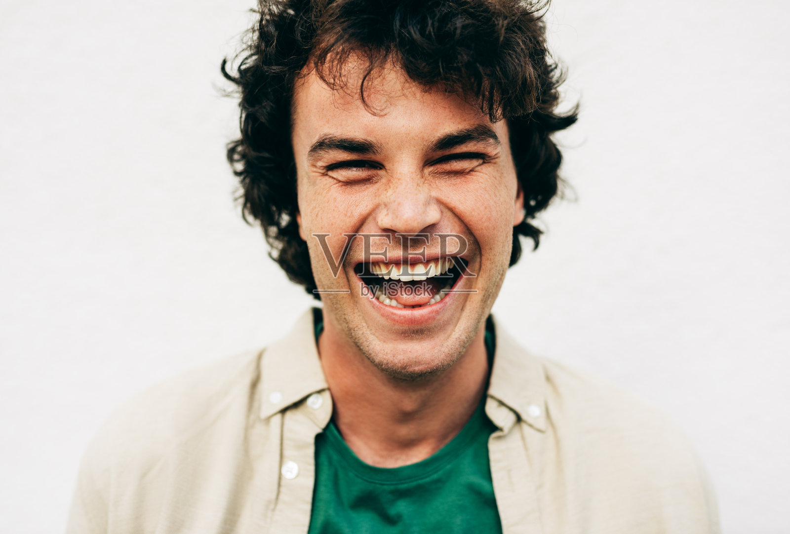 这是一个为广告摆姿势的愉快的年轻人的特写，他面带微笑，看着户外混凝土墙上的摄像机。肖像快乐聪明的学生男性卷头发微笑照片摄影图片