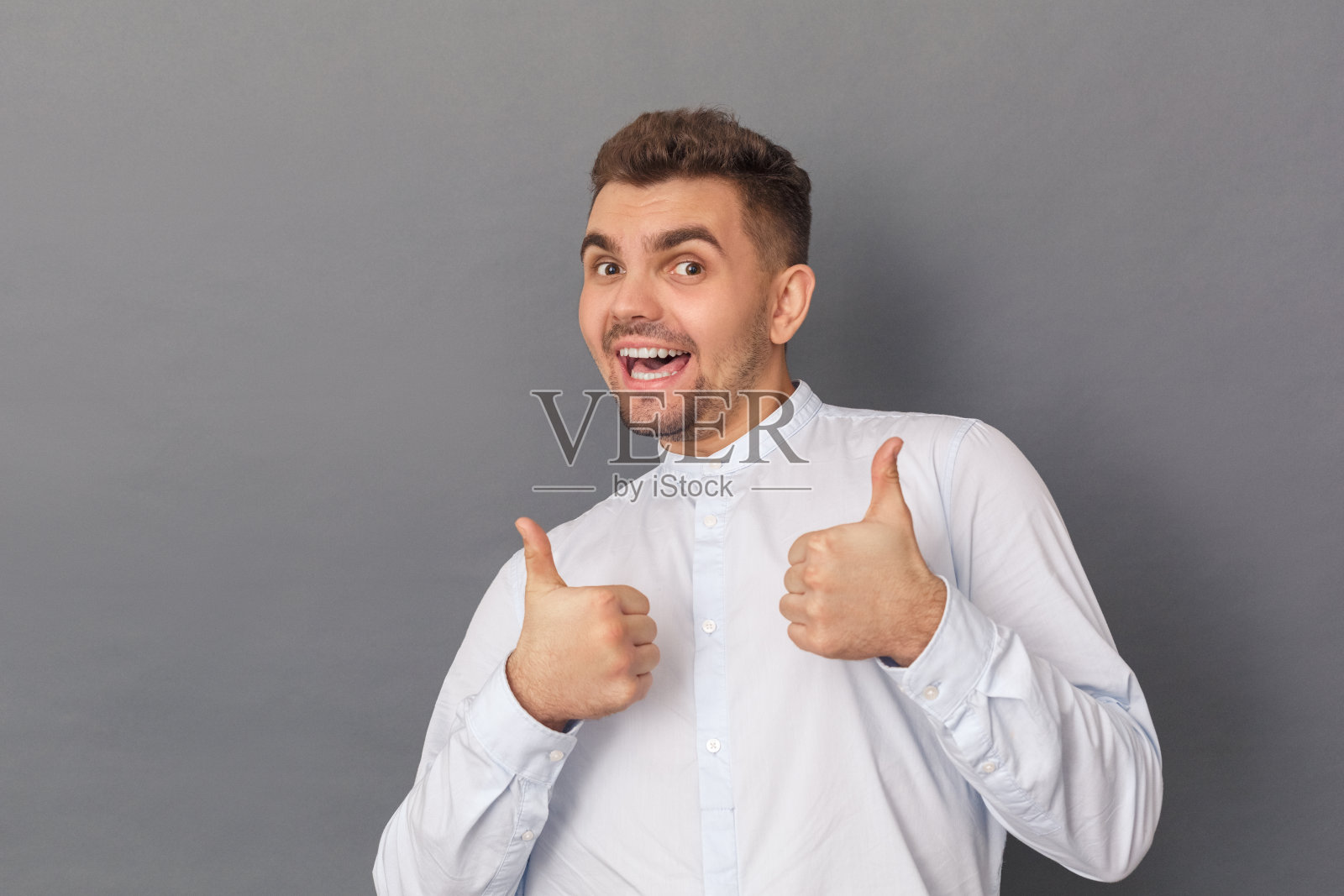 自由泳。年轻人孤立地站在灰色的工作室里，翘起大拇指，笑嘻嘻的照片摄影图片