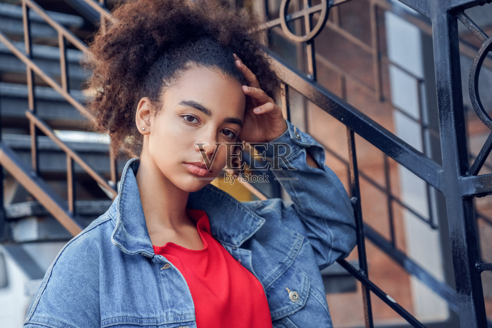 户外休闲。年轻时尚的非洲女孩在牛仔夹克站在楼梯附近触摸头部凉爽的特写镜头照片摄影图片