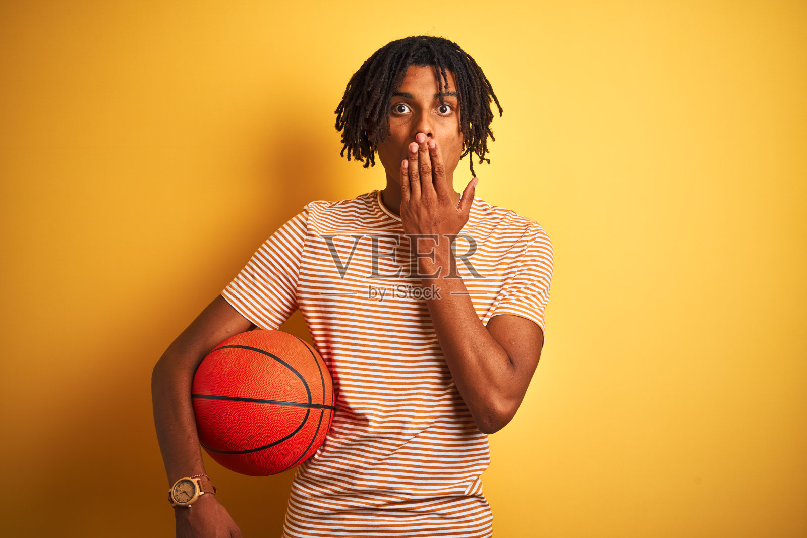 留着雷鬼辫的黑人运动员拿着篮球挡在孤立的黄色背景上，用手捂着嘴，因为错误而感到震惊，表情恐惧，沉默地害怕，秘密的概念照片摄影图片