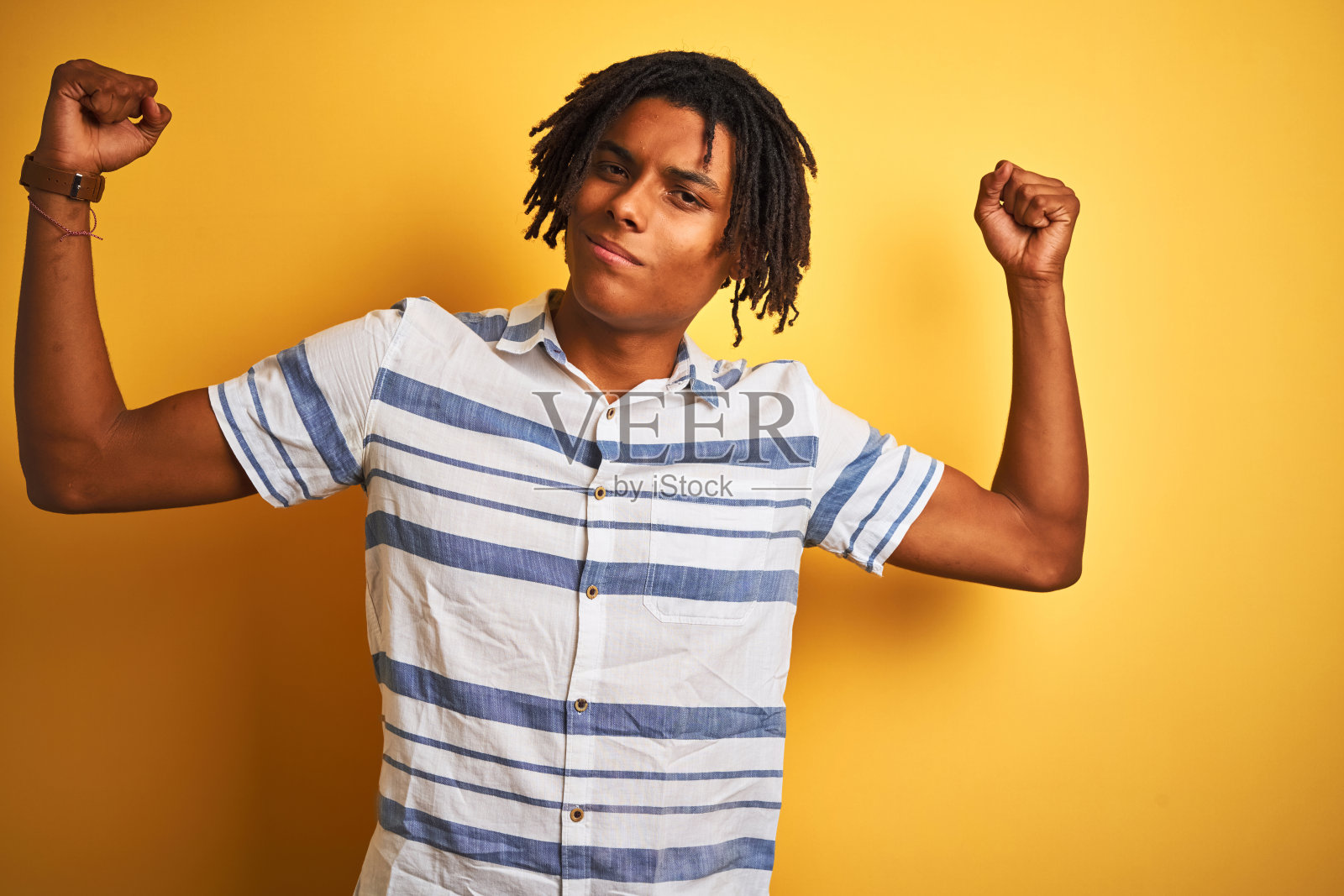 非洲裔美国人与雷鬼发穿着条纹衬衫在孤立的黄色背景显示手臂肌肉微笑自豪。健康的概念。照片摄影图片