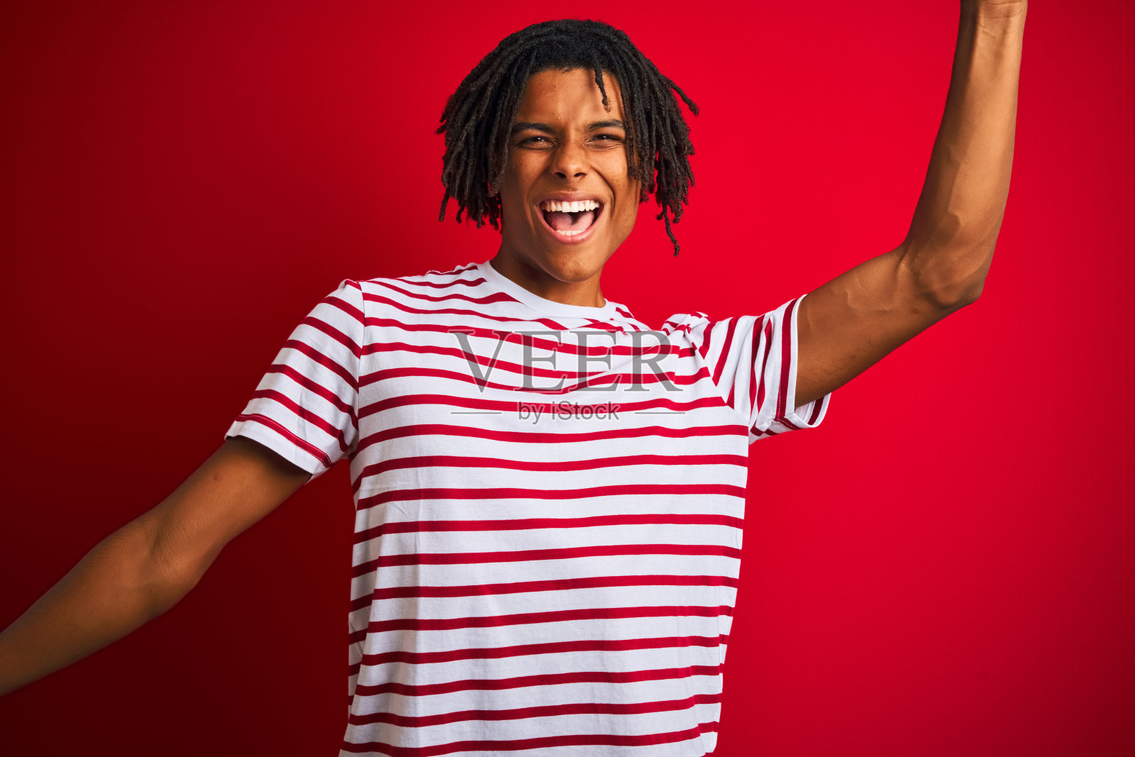 年轻的黑人，梳着雷鬼辫，穿着条纹t恤，站在孤立的红色背景上，欢快地跳舞，微笑着，听着音乐，随意而自信照片摄影图片