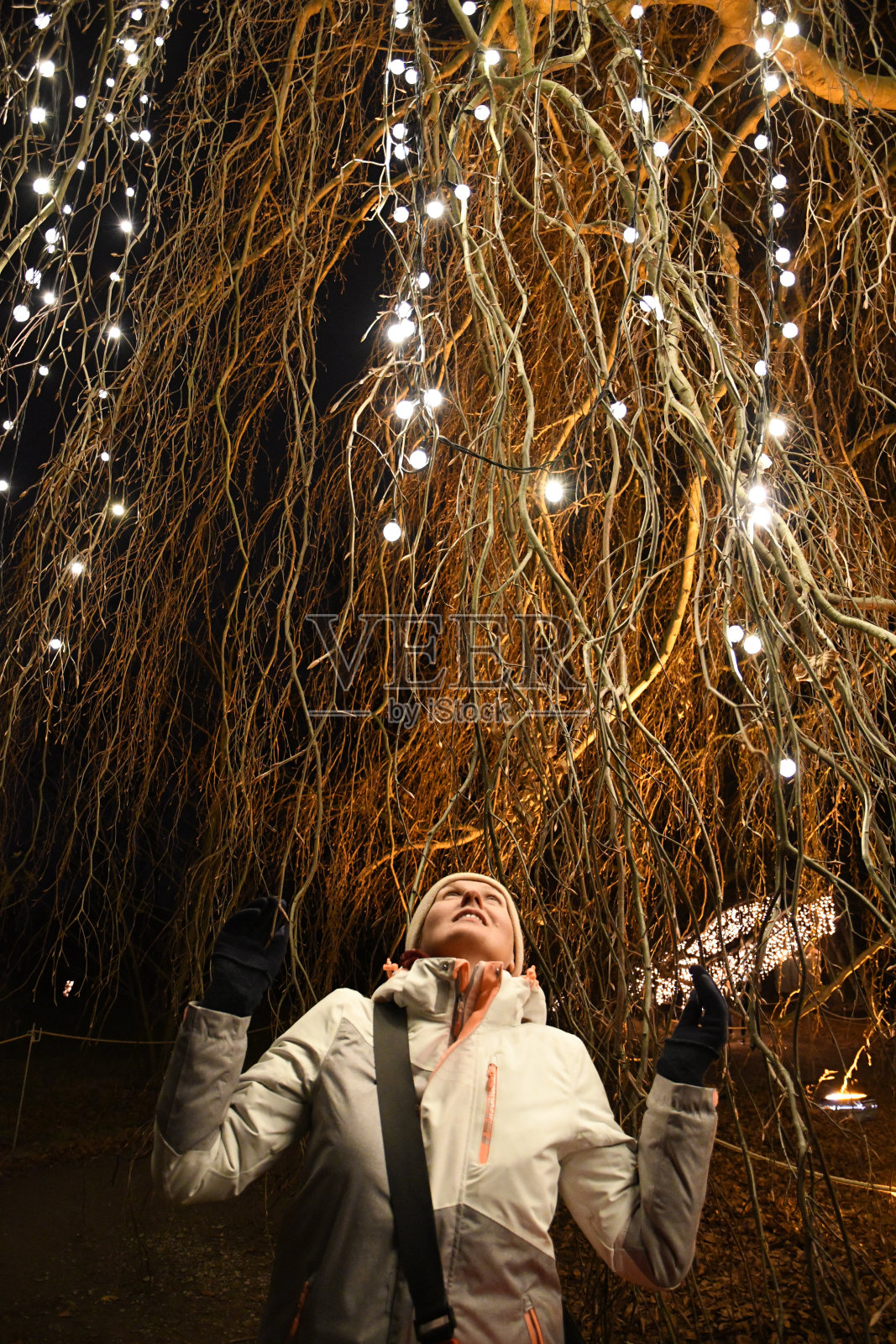 年轻女子穿着白色夹克，悬挂在巨大的柳树的树枝下，装饰着闪闪发光的圣诞灯和花环，在夜晚的黑暗中闪闪发光。神奇的树枝照明。节日场景照片摄影图片