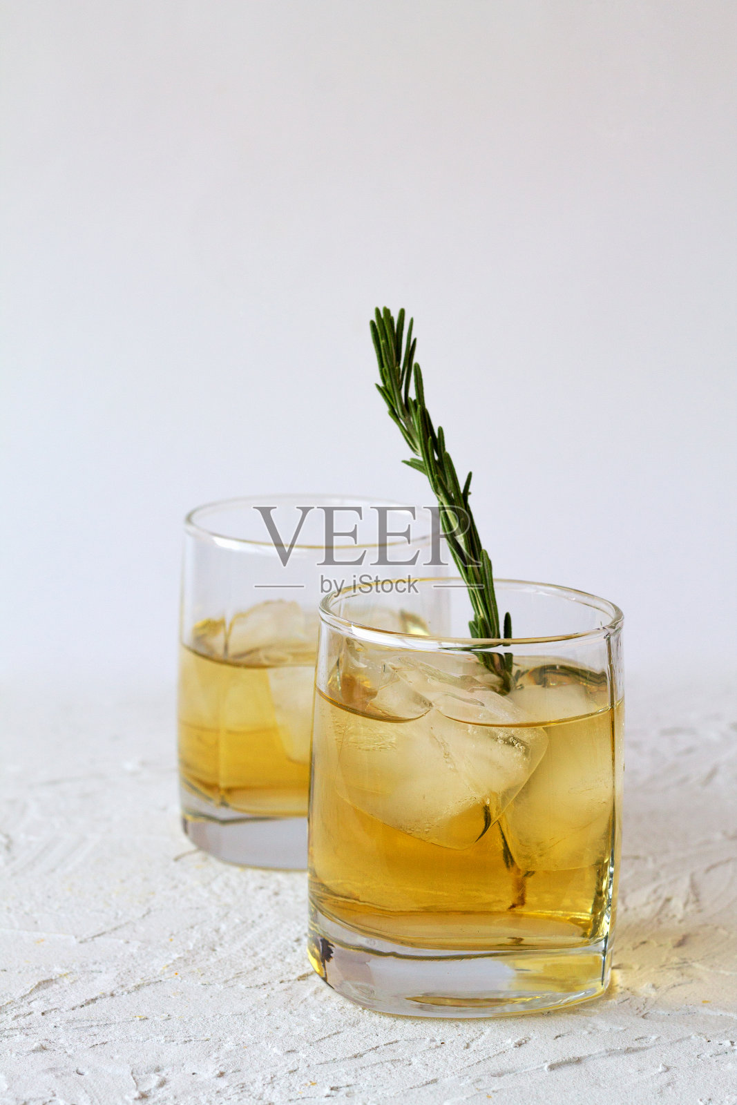 一杯加冰块的柠檬水，在白色的背景上放一小枝迷迭香。白桌子上放两杯加冰的威士忌。照片摄影图片