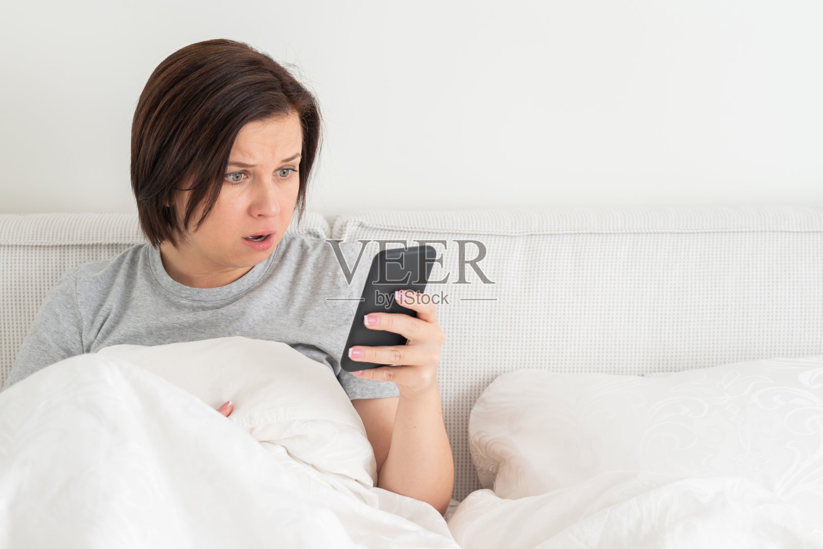 一个穿着羽绒被坐在床上玩智能手机的惊讶女人照片摄影图片