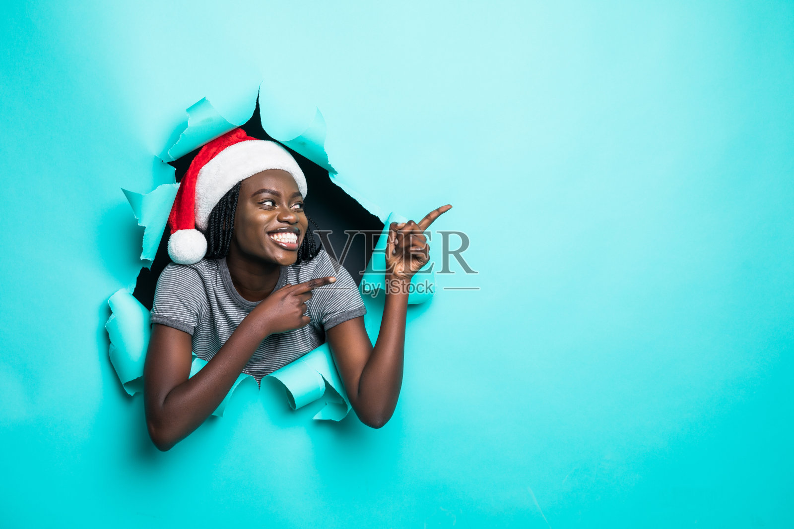 年轻的非洲妇女戴在圣诞老人的帽子指向与手在复印空间站在绿色撕纸洞的背景照片摄影图片