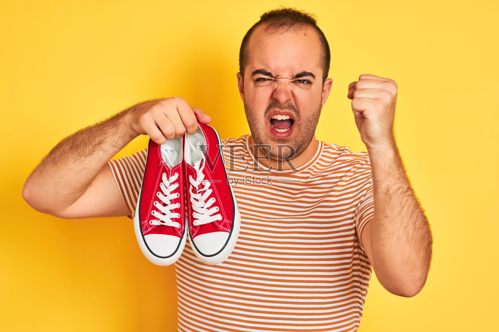 年轻人拿着红色的休闲运动鞋站在孤立的黄色背景恼怒和沮丧的呼喊与愤怒，疯狂的呼喊与举起的手，愤怒的概念照片摄影图片
