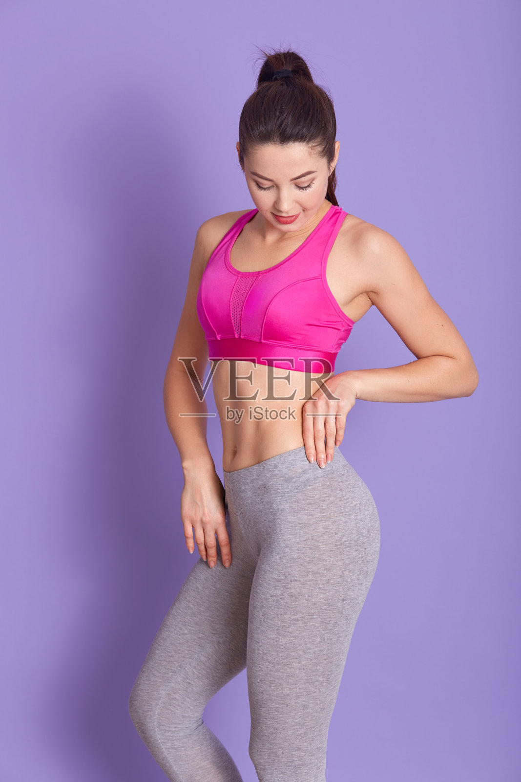 全长照片的运动良好的运动员孤立在工作室的淡紫色背景，低头，穿着灰色leggins和粉色上衣，有马尾辫。人与健身理念。照片摄影图片