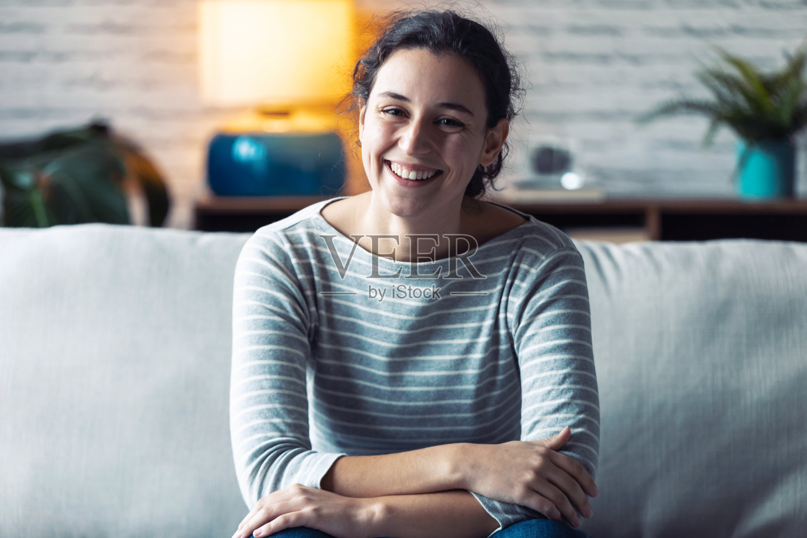 微笑的年轻女子看着相机，坐在沙发上，在家里的客厅。照片摄影图片