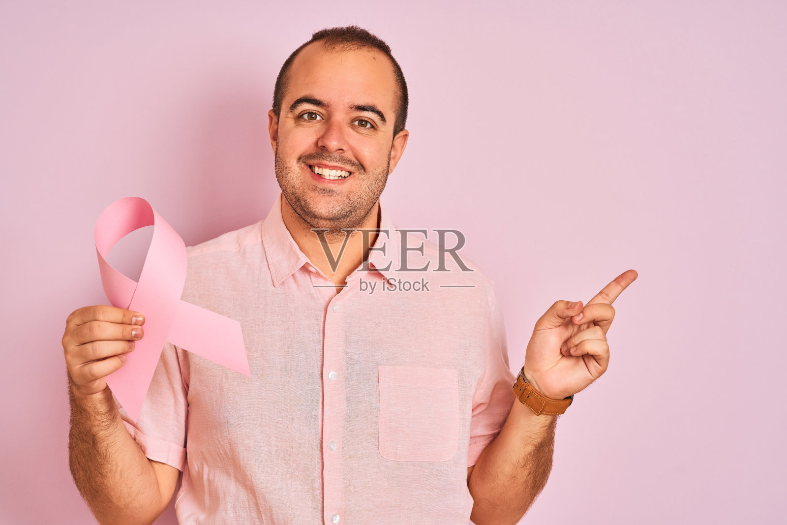 年轻男子举着癌症丝带标志站在孤立的粉红色背景上，非常高兴地用手指向一边照片摄影图片