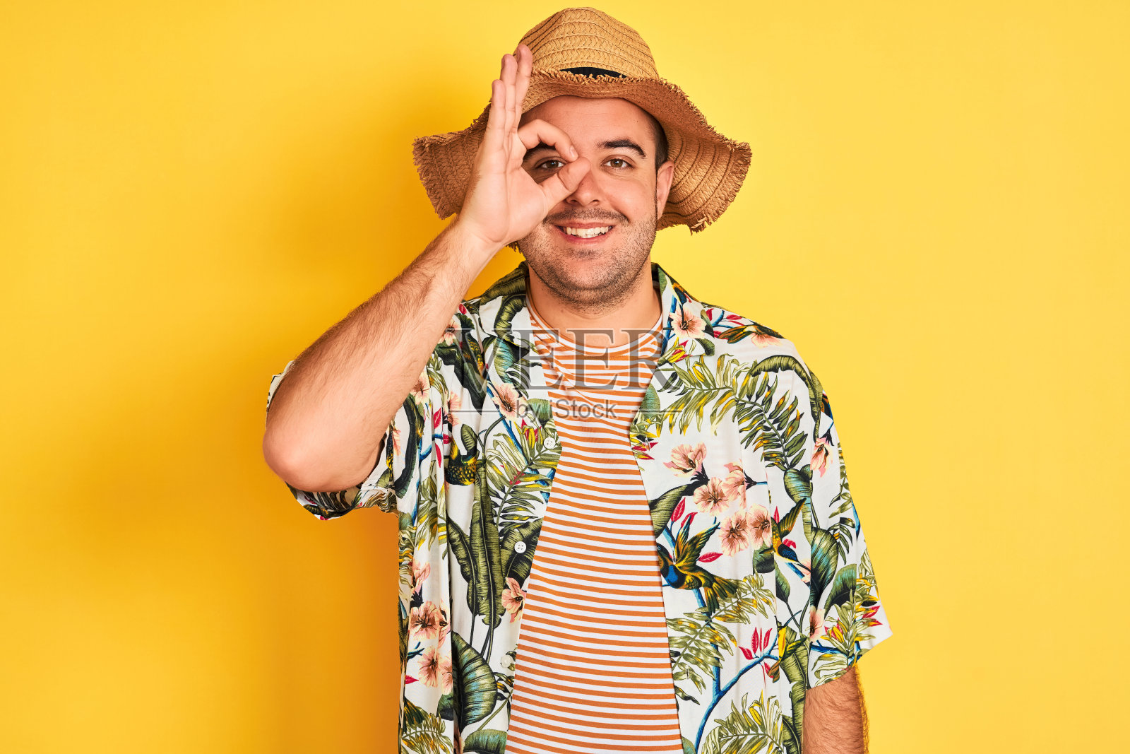 年轻男子度假穿夏季衬衫和帽子在孤立的黄色背景做ok手势与手微笑，眼睛透过手指与幸福的脸。照片摄影图片