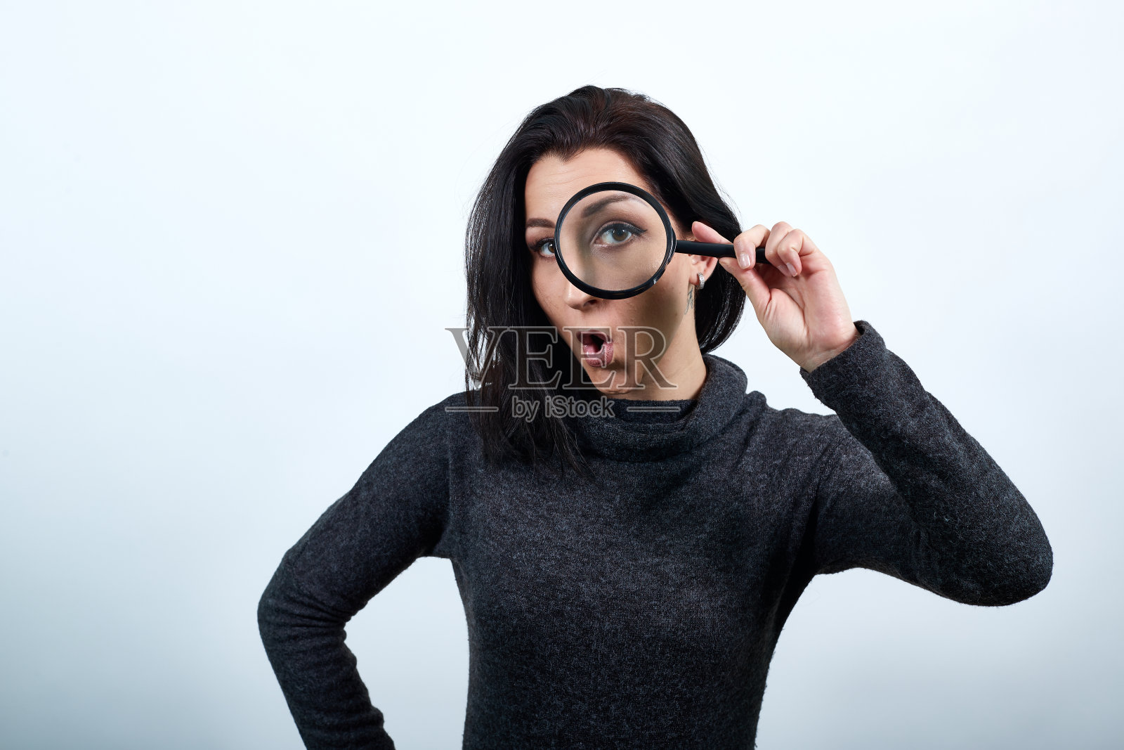 震惊的年轻女子把放大镜放在眼睛附近，手放在腰上照片摄影图片