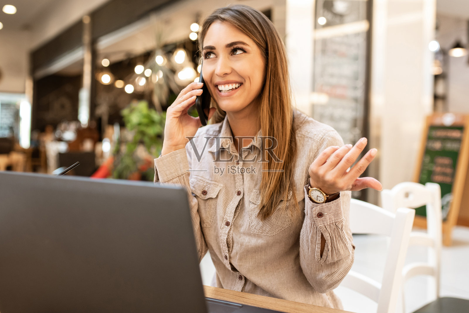 美丽的年轻褐发女人微笑着和智能手机上的朋友聊天，笔记本电脑在她面前的咖啡厅。照片摄影图片