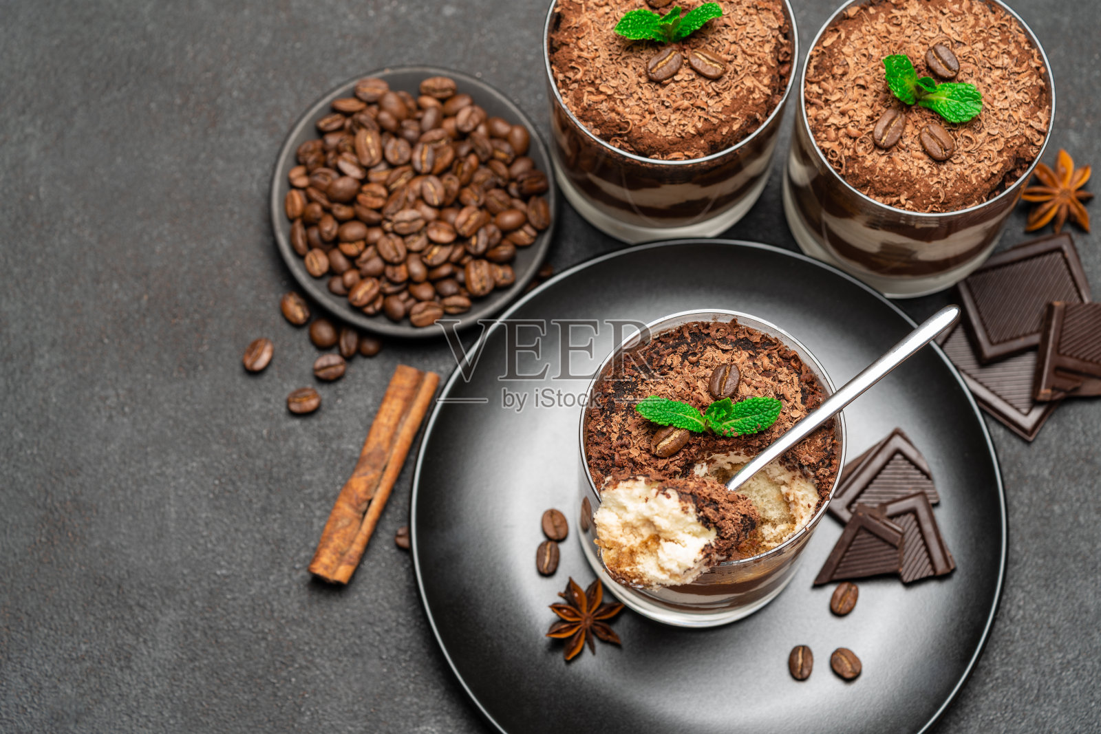 经典的提拉米苏甜点装在玻璃杯里，黑色混凝土背景下的石头切板上有几块巧克力照片摄影图片