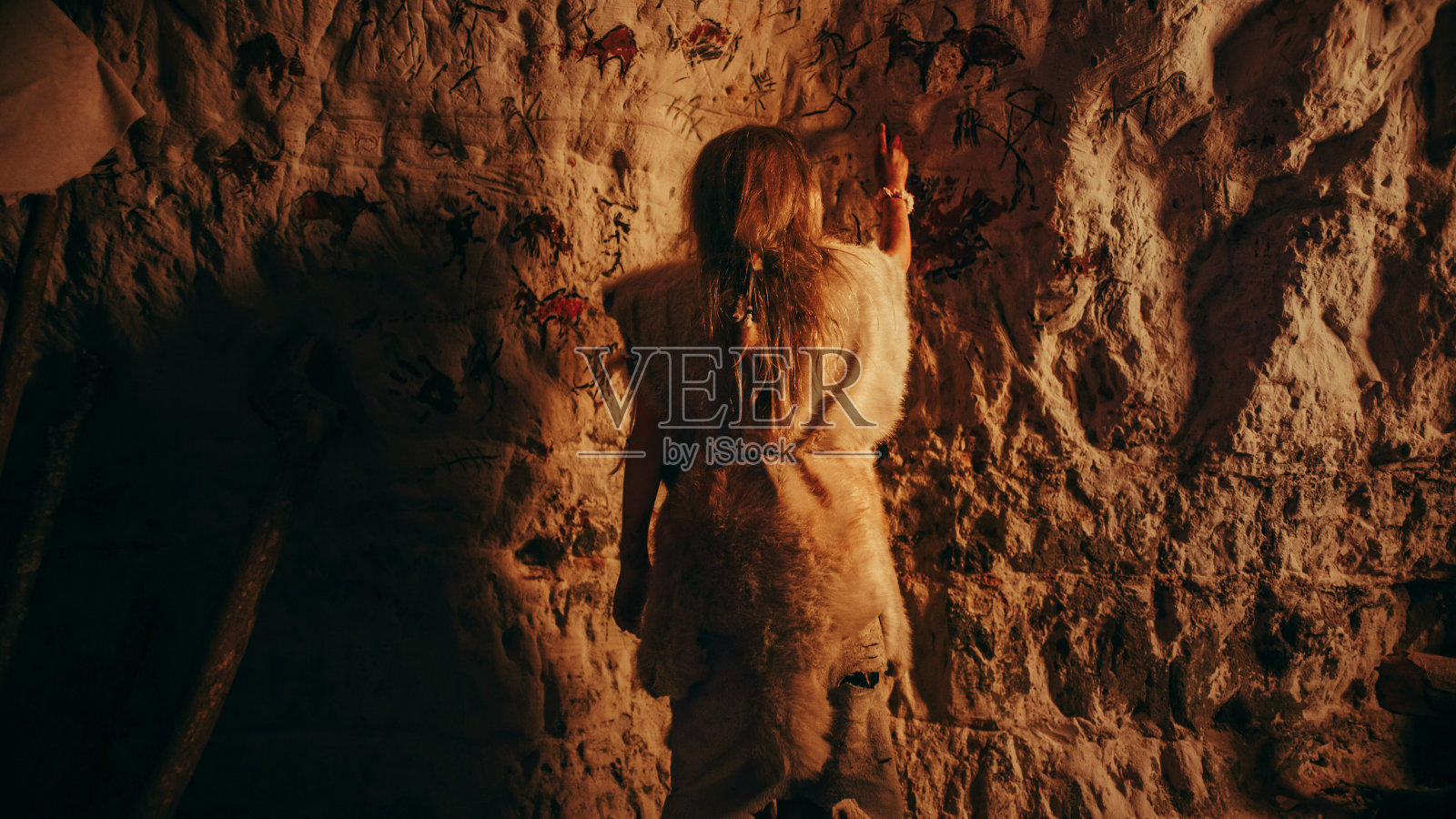 一个原始史前尼安德特人的孩子穿着动物的皮肤在晚上的墙壁上画动物和抽象。创造第一个岩画洞穴艺术。照片摄影图片