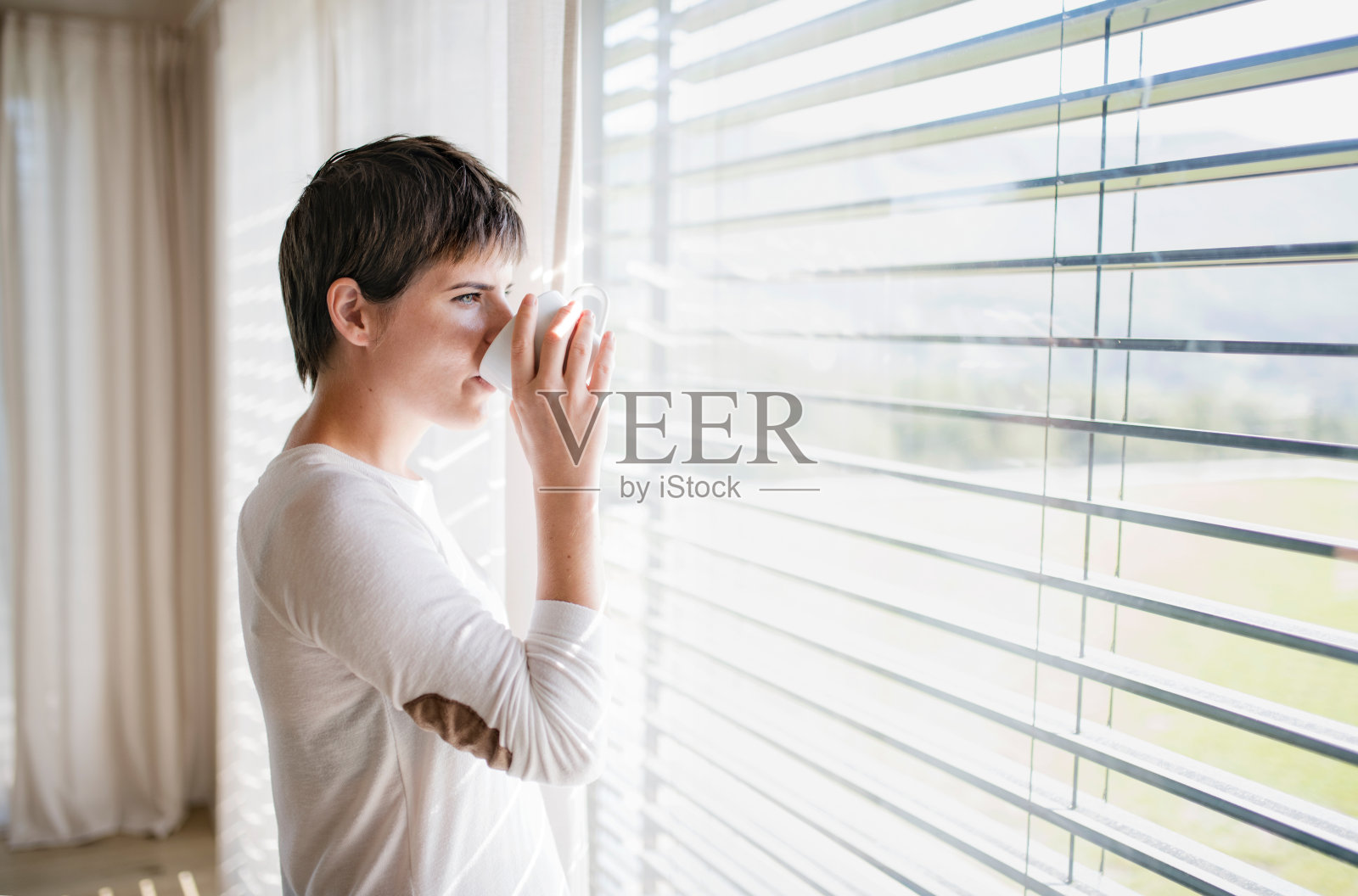 年轻女子的肖像，咖啡站在家里的窗边。照片摄影图片