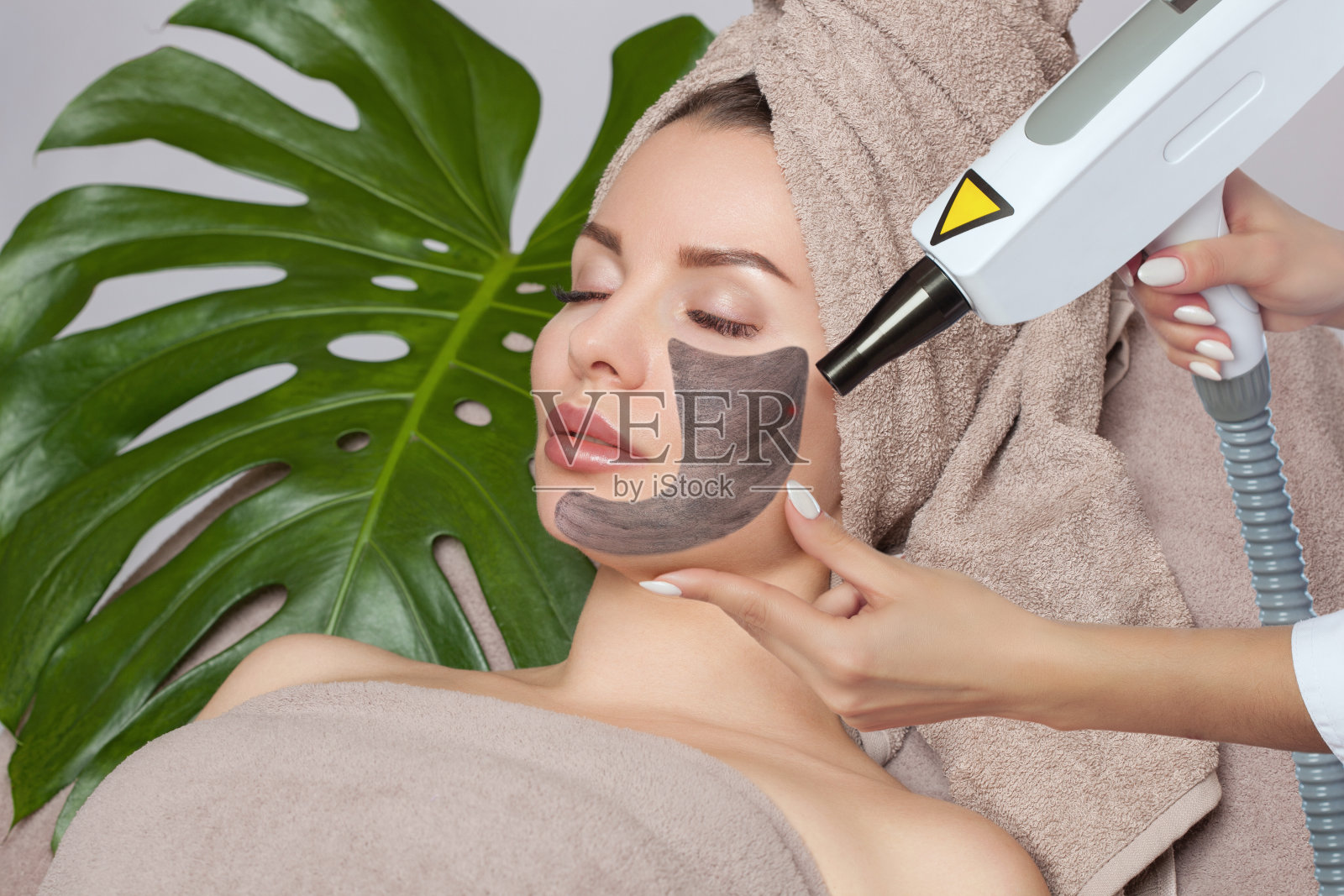 一个脸上戴着碳面膜的女人，美容师在年轻美丽的女孩皮肤有问题的地方做激光碳剥离。附近有一片怪物的叶子。硬件美容。照片摄影图片