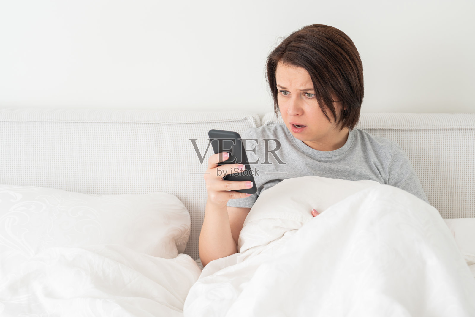 一个穿着羽绒被坐在床上玩智能手机的惊讶女人照片摄影图片