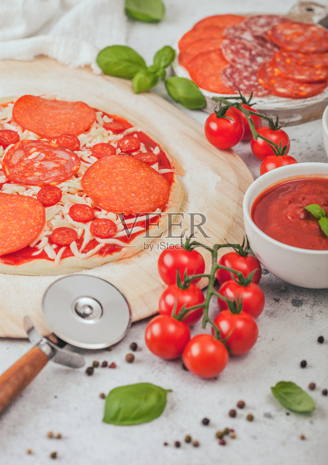 准备烤意大利辣香肠披萨，用滚轮切刀，新鲜的番茄和罗勒，配上带有奶酪和番茄酱的碗盘。照片摄影图片