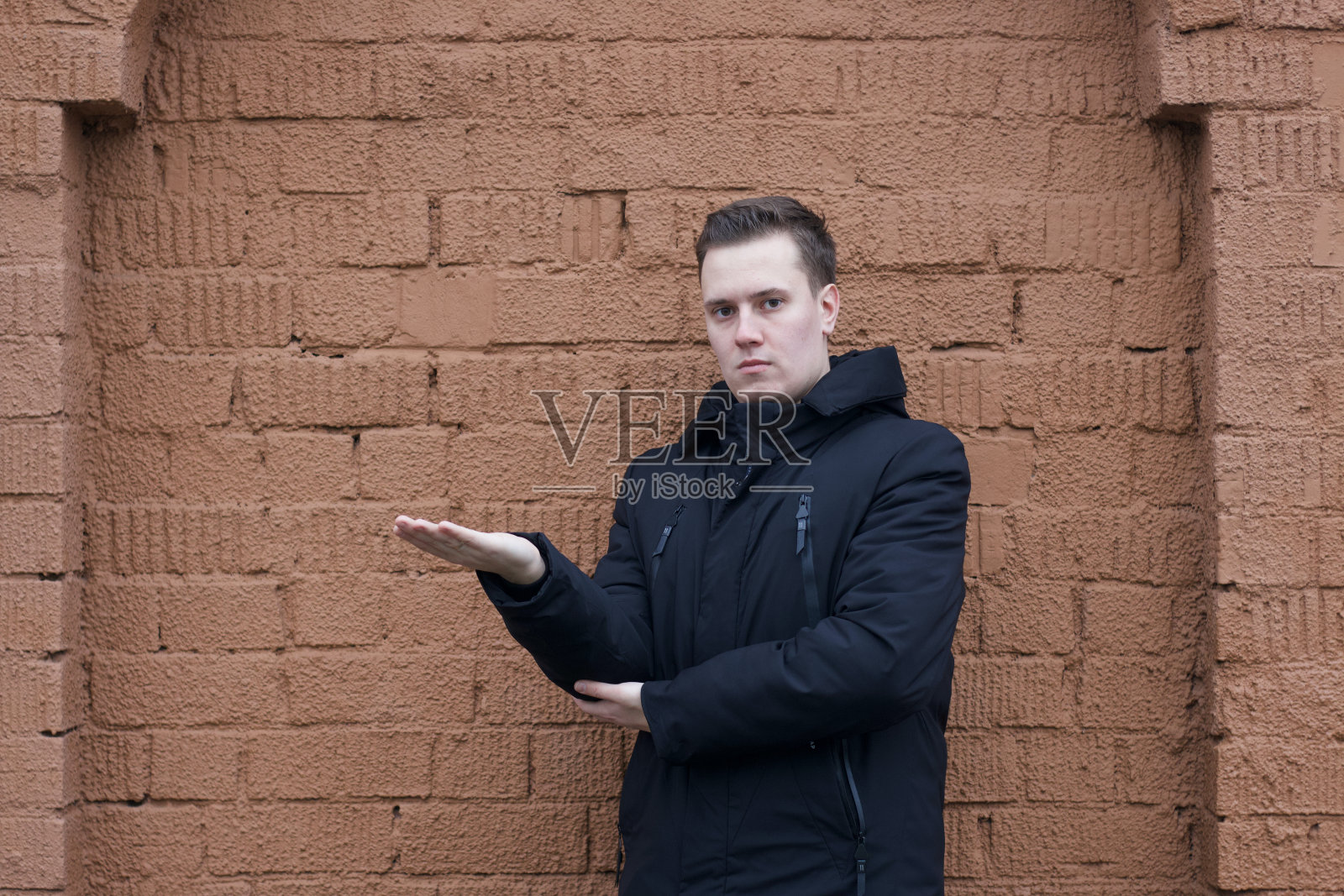 一名身穿黑色夹克的年轻人在一堵棕色砖墙的背景上拍照。他把手放在一边，掌心朝上。照片摄影图片