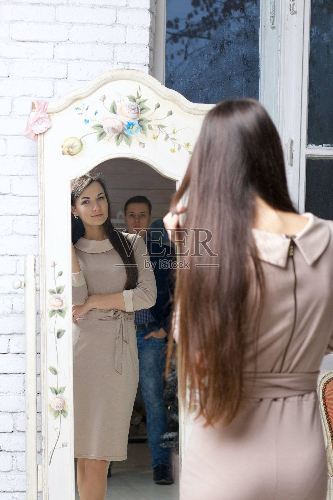 镜子里的女孩。在她的肩膀后面是正在看她的男人的影子。那个女孩穿着一件米色的连衣裙。地板上放着一面老式的镜子。照片摄影图片