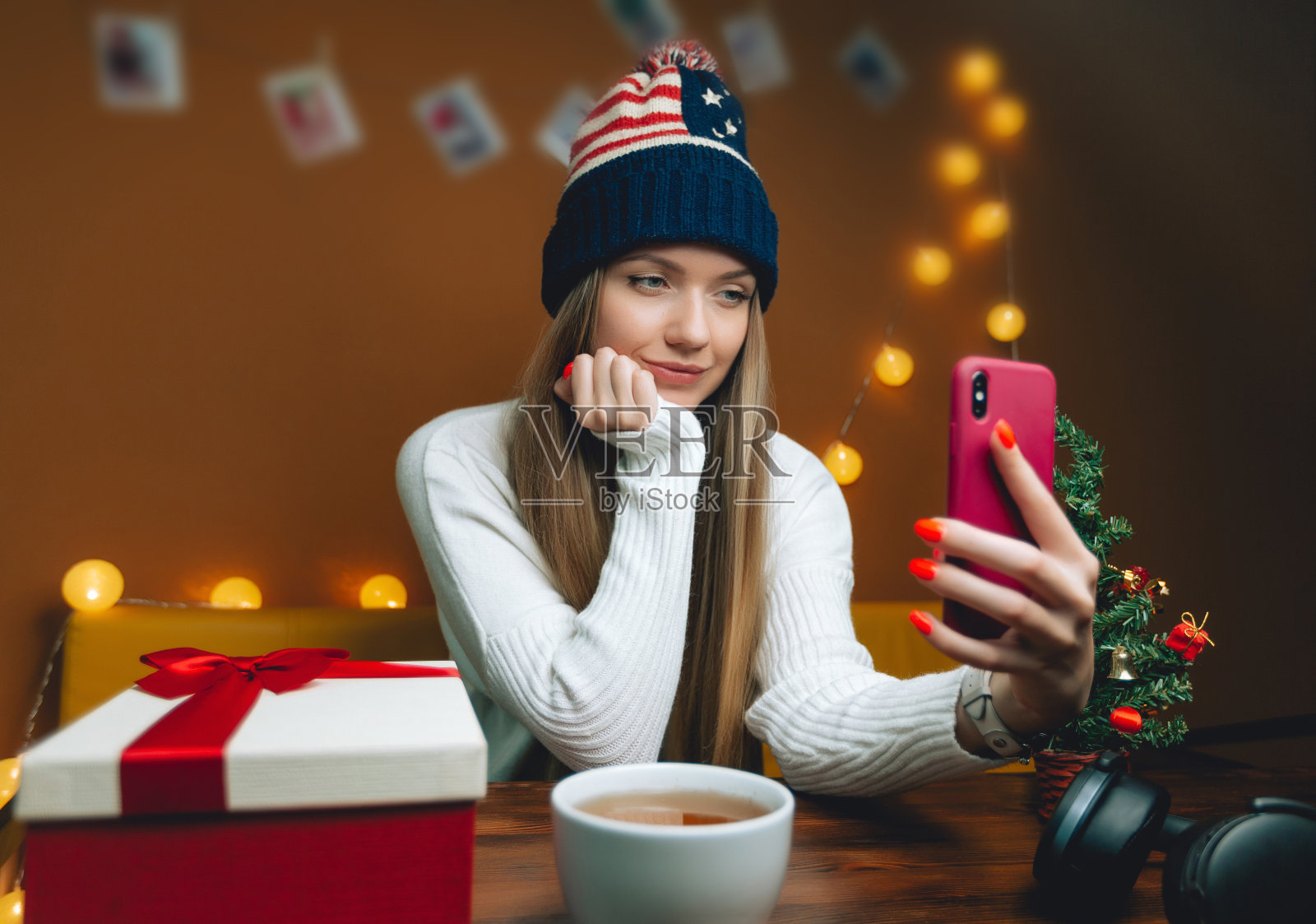 戴着美国帽子的漂亮女人在用智能手机自拍。圣诞树放在木桌上照片摄影图片