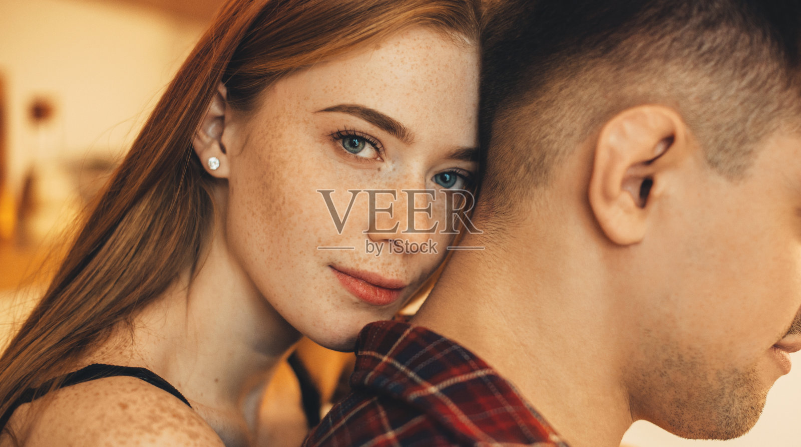 美丽的年轻女性与红头发和雀斑看着镜头微笑，而从背后拥抱她的男友在厨房。照片摄影图片