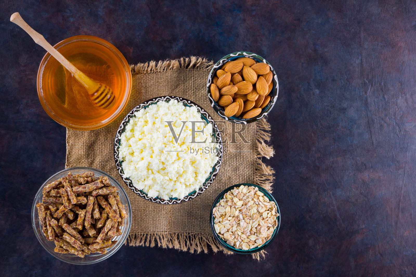 健康饮食早餐，粗麻布。健康早餐的配料-燕麦片，格兰诺拉麦片，蜂蜜，杏仁，软干酪。深色背景上自制的格兰诺拉麦片。乡村风格照片摄影图片