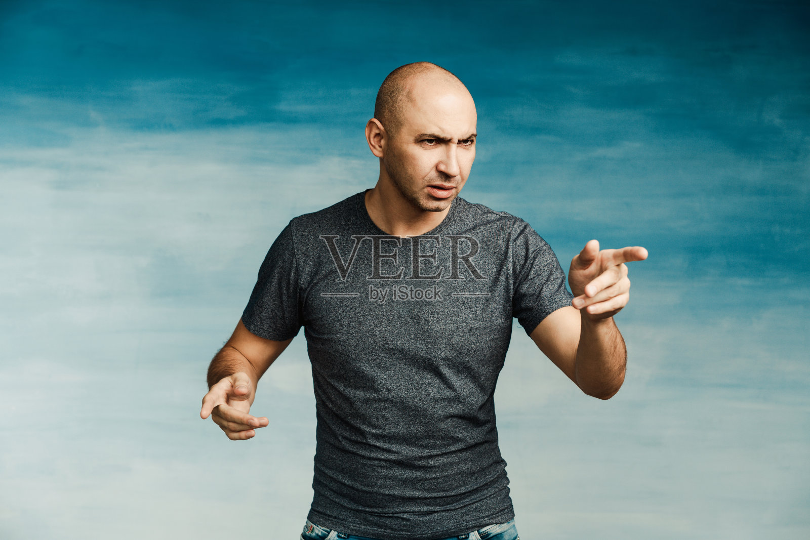 一个穿着灰色t恤和牛仔裤的秃顶年轻男子恶意地用手指着一边照片摄影图片