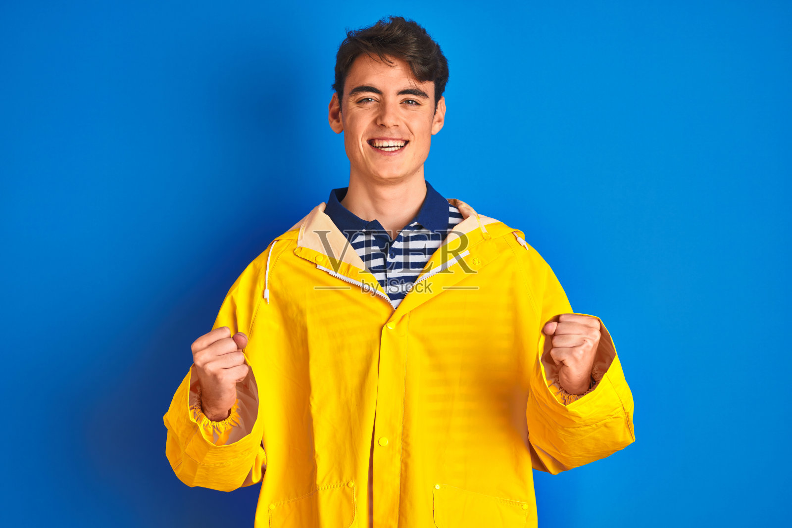 十几岁的渔民男孩穿着黄色雨衣在孤立的背景庆祝惊讶和惊讶的成功，举起手臂和睁开眼睛。赢家的概念。照片摄影图片