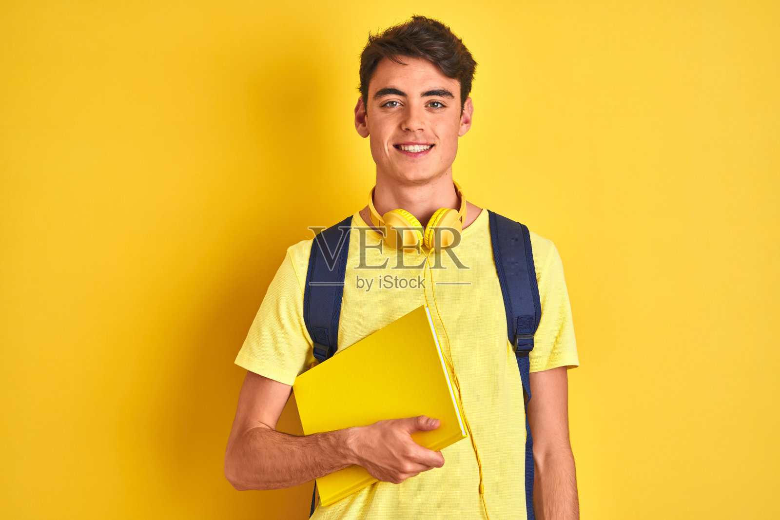 十几岁的男孩戴着耳机和背包在孤立的背景阅读一本书，一个快乐的脸站着，自信的微笑露出牙齿照片摄影图片