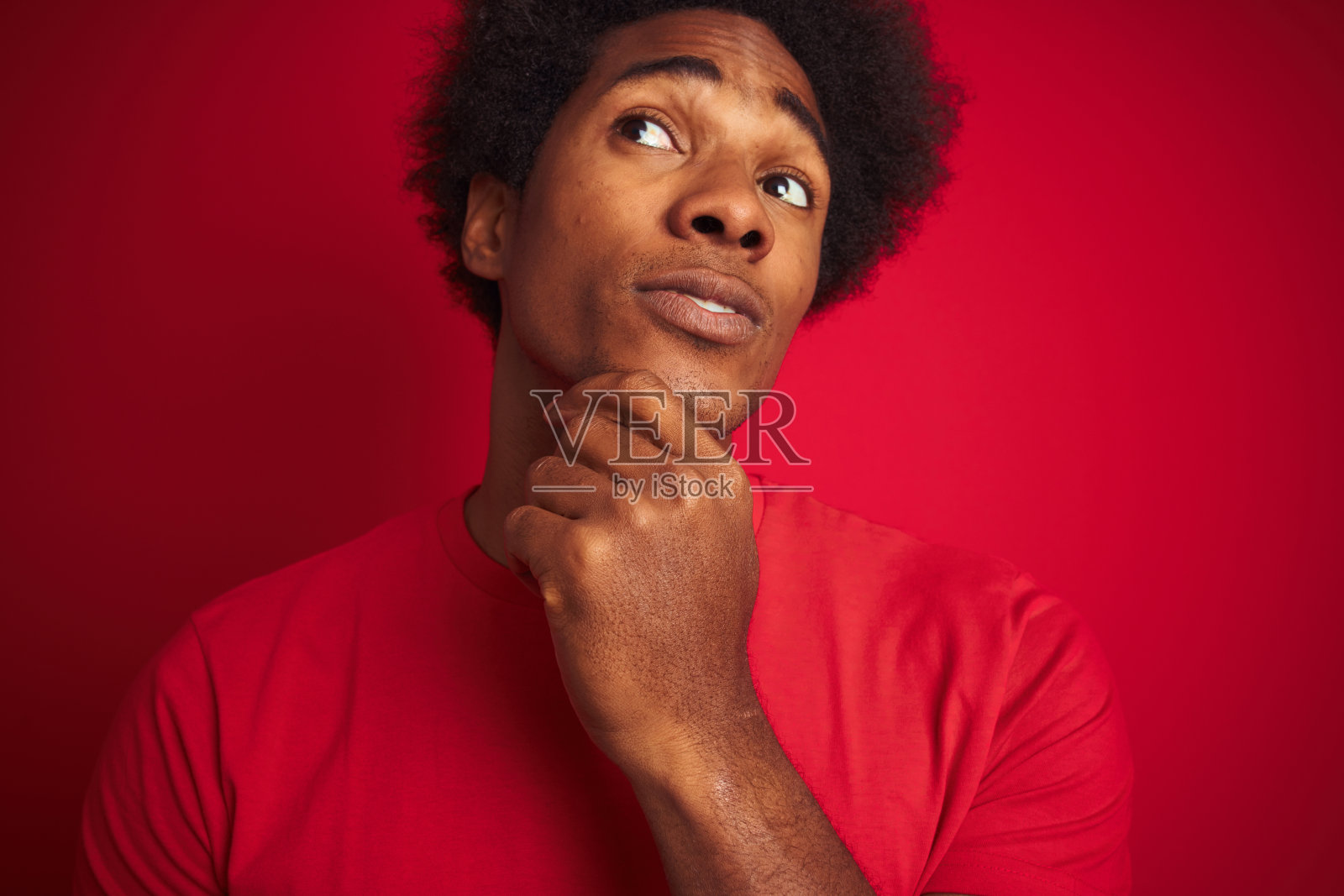 一个留着非洲式发型穿着t恤的美国青年站在孤立的红色背景严肃的脸上思考问题，非常困惑的想法照片摄影图片
