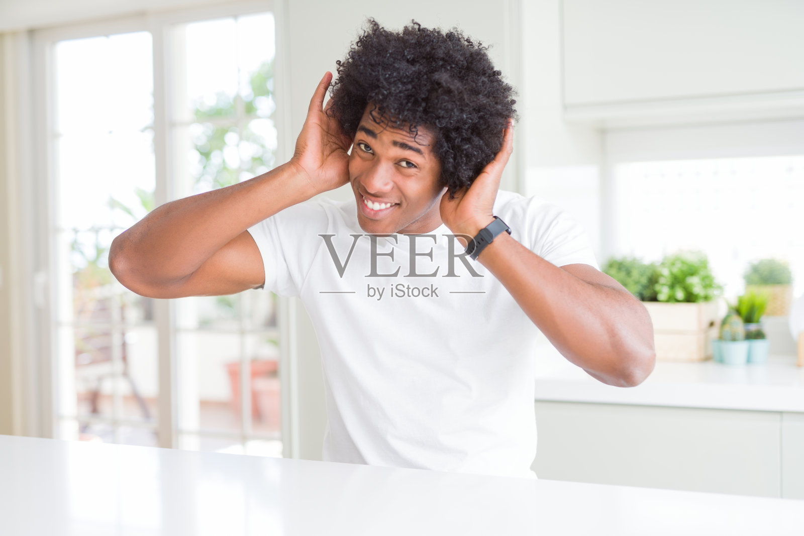 年轻的非洲裔美国人穿着休闲的白色t恤坐在家里试图听到双手在耳朵上的手势，好奇的八卦。听力问题,充耳不闻照片摄影图片