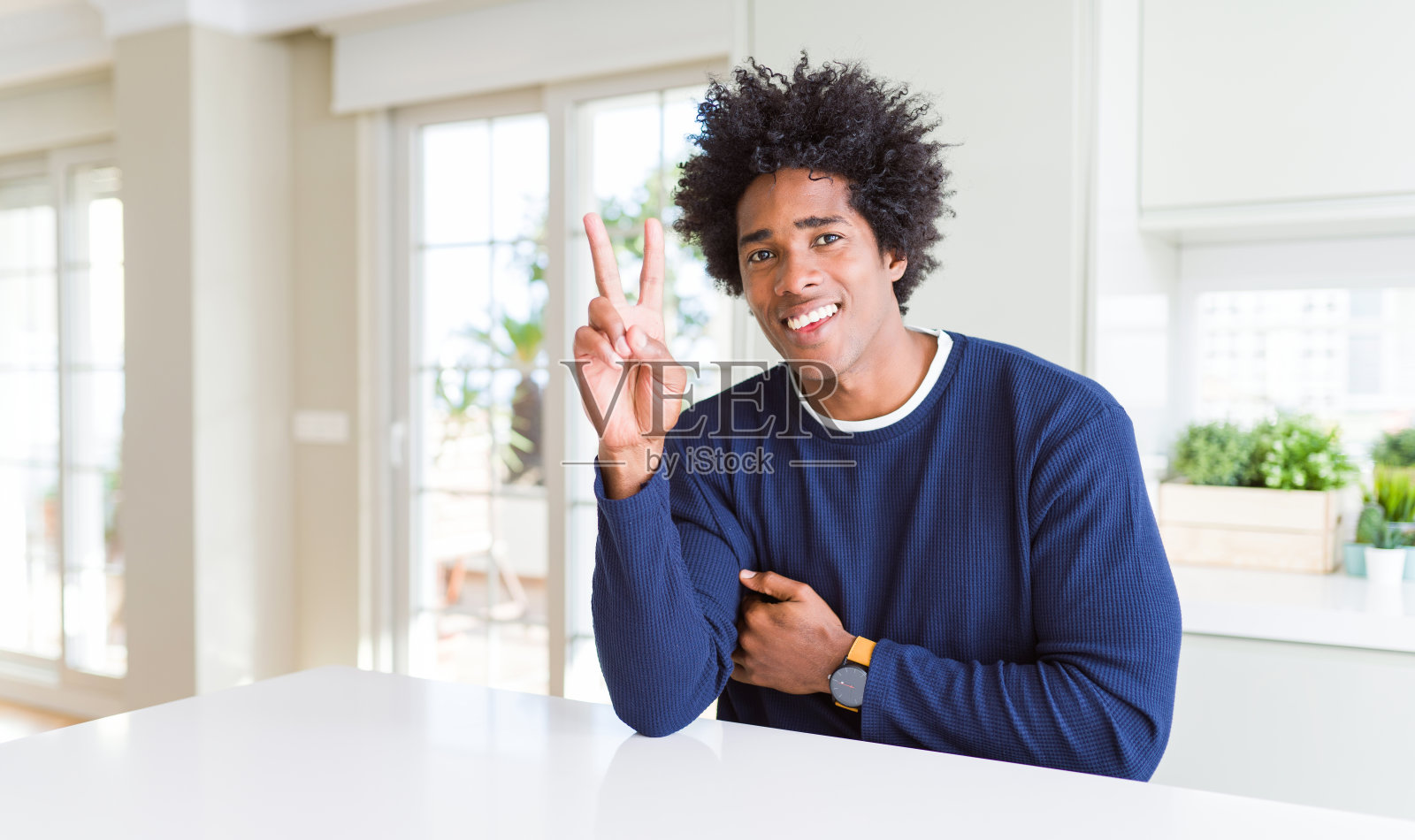 年轻的非洲裔美国人穿着休闲毛衣坐在家里微笑着看着摄像机显示手指做胜利手势。二号人物。照片摄影图片